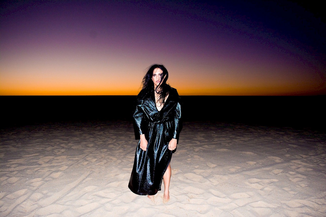 ALKANARA - Diego Bragà is in the center of the photograph, looking directly into the lens, with arms hanging by their side. They are dressed in a long, shiny black coat, reaching down to their bare feet buried in the sand. In the background, the horizon line and the evening sky, fading from orange to purple. - ©Rui Palma