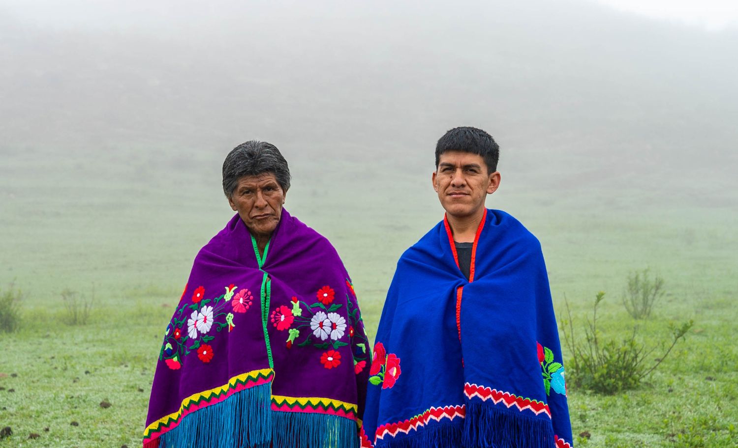 Vemos duas pessoas à frente de uma paisagem de natureza, verde, com nevoeiro. Essas pessoas usam trajes tradicionais e coloridos e olham para a frente, fitando a câmara. - Matias Gutierrez