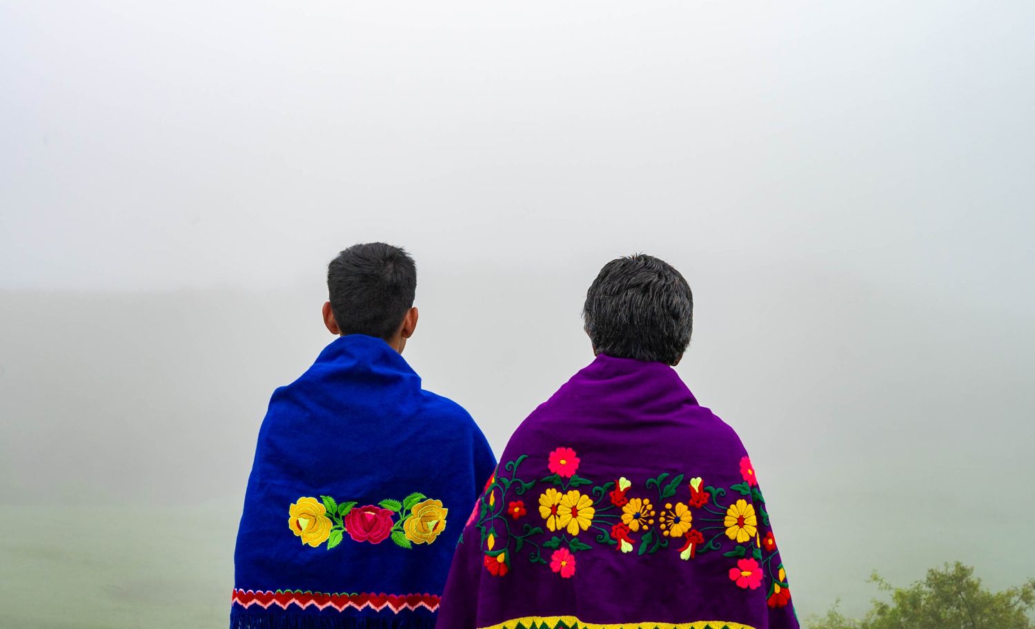 Vemos duas pessoas, de costas, com uma paisagem com nevoeiro ao fundo. As pessoas usam trajes tradicionais coloridos. - Matias Gutierrez