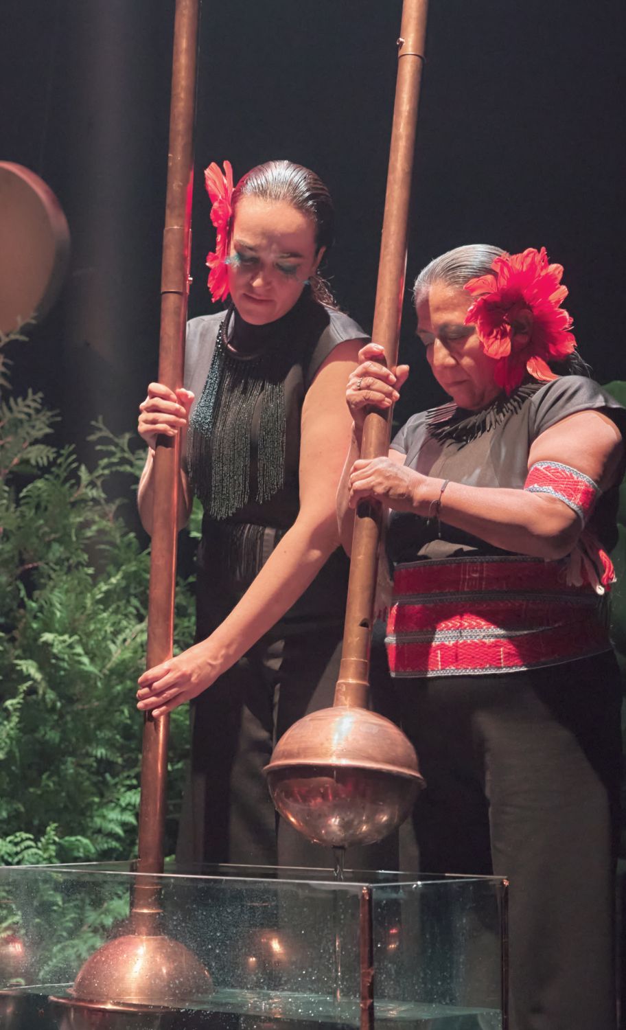 Two people are standing holding two large copper-coloured objects. They have a small tank of water in front of them, where one of the objects is inserted and the other has just come out. The people are dressed in black and red, looking down with an air of concentration. On the left-hand side we see a green plant in the background. - Antoine Raymond