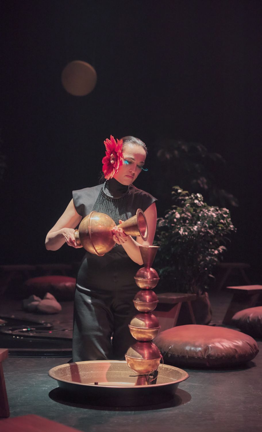 A person is kneeling on the floor and holding a large, copper-coloured object, leaning towards another object. The person is dressed in black, has a red flower attached to the face and large green eyebrows. - Antoine Raymond