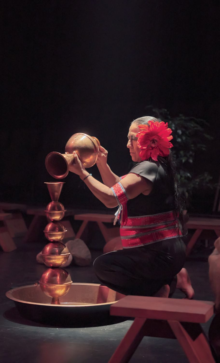 A person has their knees on the floor and is holding a large, copper-coloured object, tilting it towards another object. We see the person in profile. The person is dressed in black and red and has a red flower attached to their face. The room is dark, and on the floor we can see several benches scattered around. - Antoine Raymond