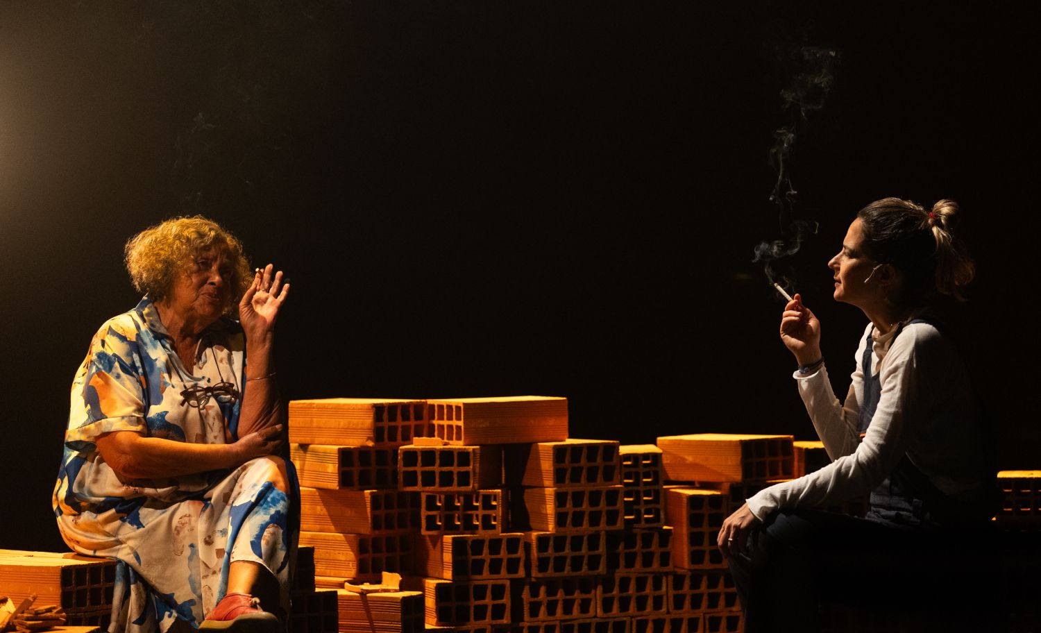 Duas pessoas estão sentadas num palco, com uma pilha de tijolos posicionada à sua frente, a conversar. A pessoa da direita está a fumar um cigarro. - Elisa Freitas
