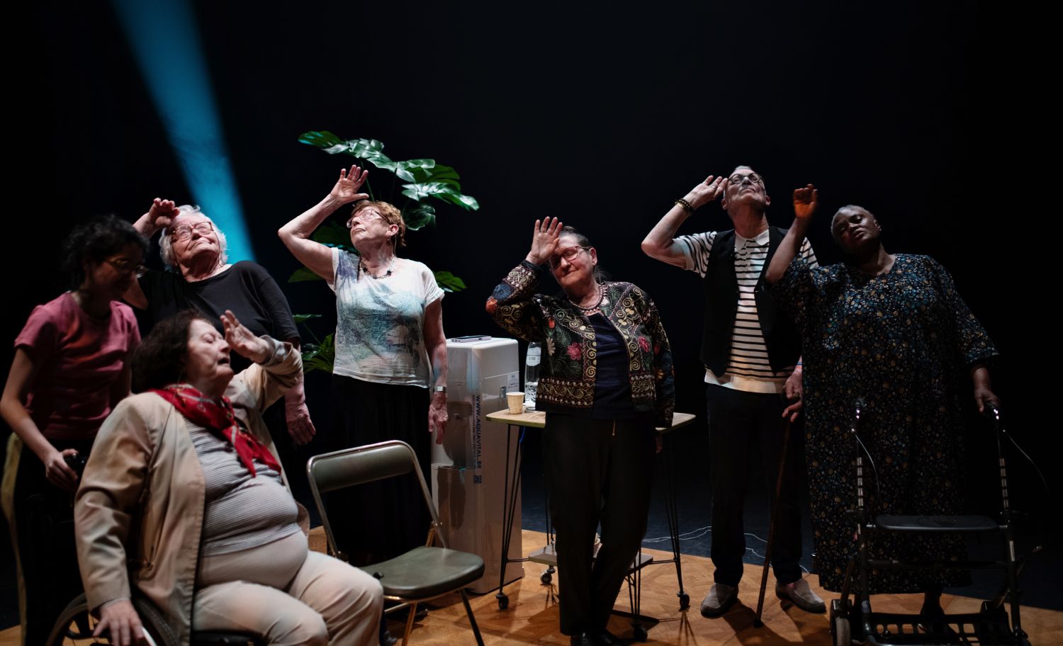 Grupo de pessoas em palco com posturas expressivas, todos com as mãos levantadas. Há um foco de luz e uma planta visível. - Yohanne Lamoulère / Tendance Floue
