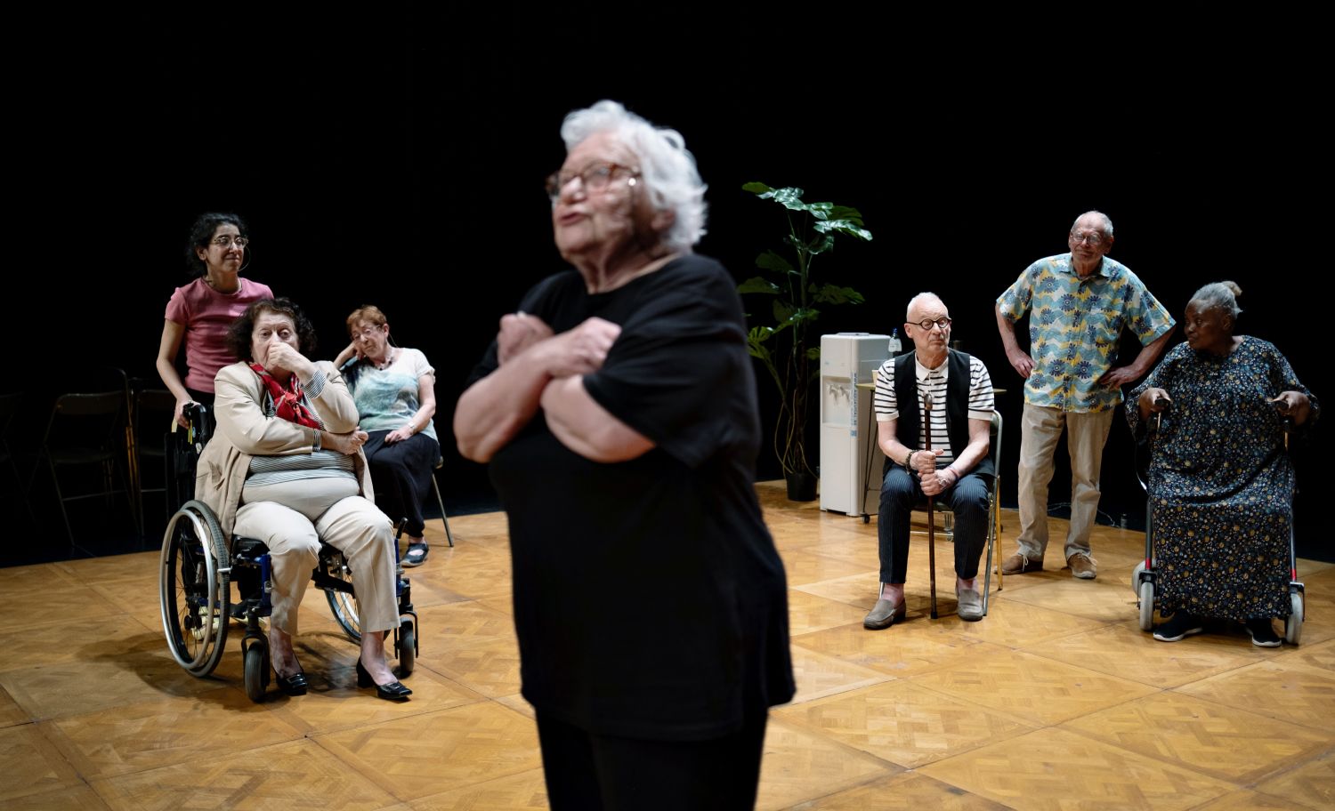 Um grupo de pessoas reunidas no palco, à volta de uma pessoa idosa que está de pé, no meio, com os braços cruzados. Há uma planta e um frigorífico ao fundo. - Yohanne Lamoulère / Tendance Floue