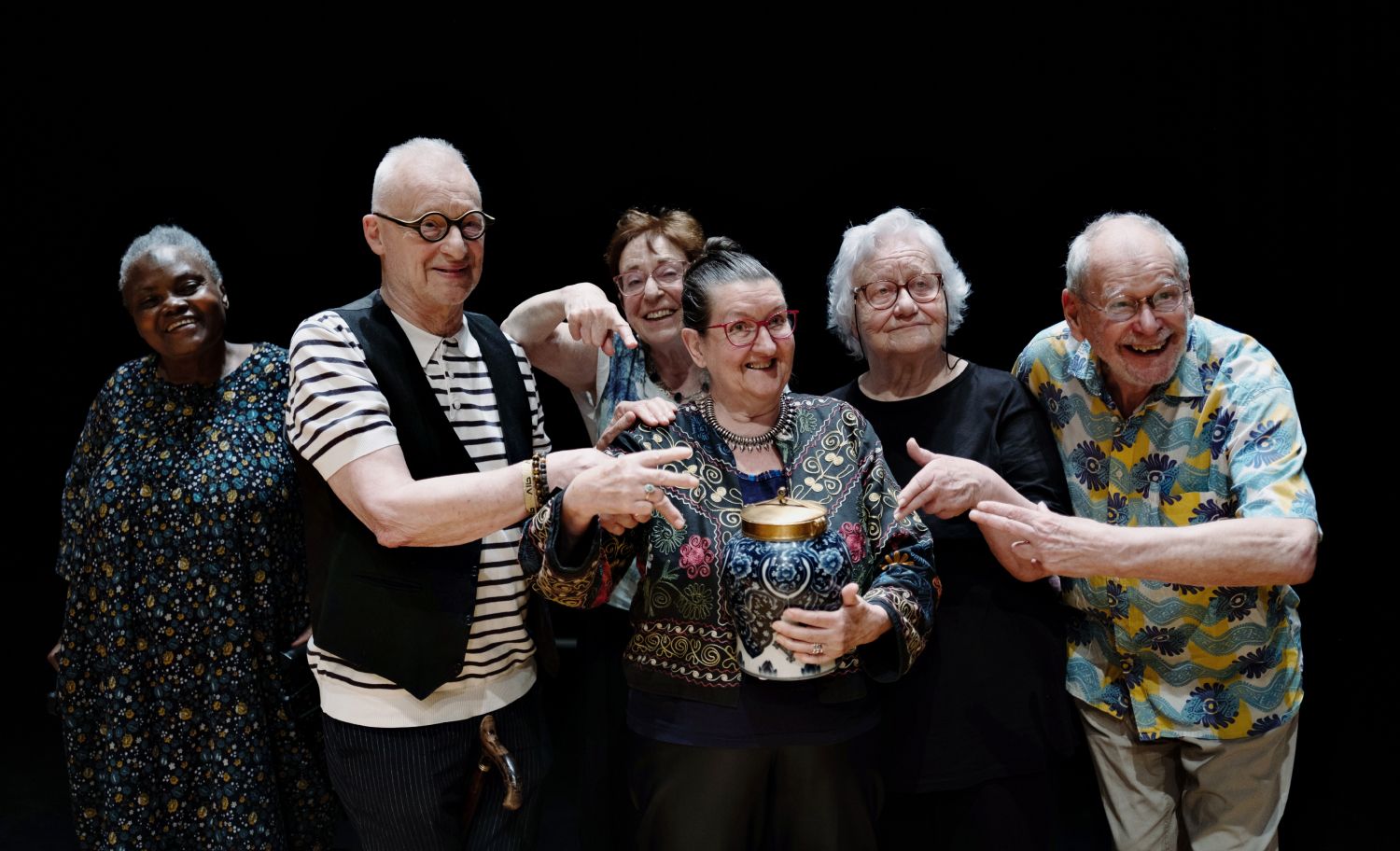 Um grupo alegre de adultos mais velhos sorri e aponta para um vaso, sobre um fundo preto. - Yohanne Lamoulère / Tendance Floue