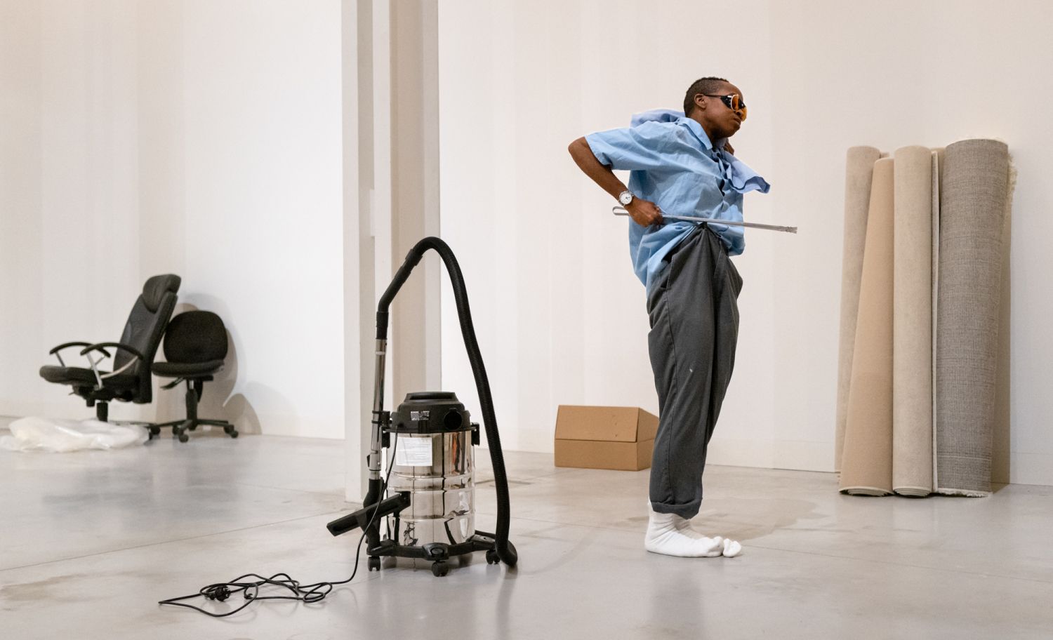 Uma pessoa está puxar as suas calçasde costas para um aspirador numa sala vazia com alguns objetos de escritório. A pessoa sem sapatos e veste uma camisa azul, calças cinzentas e meias brancas. - Françoise Robert