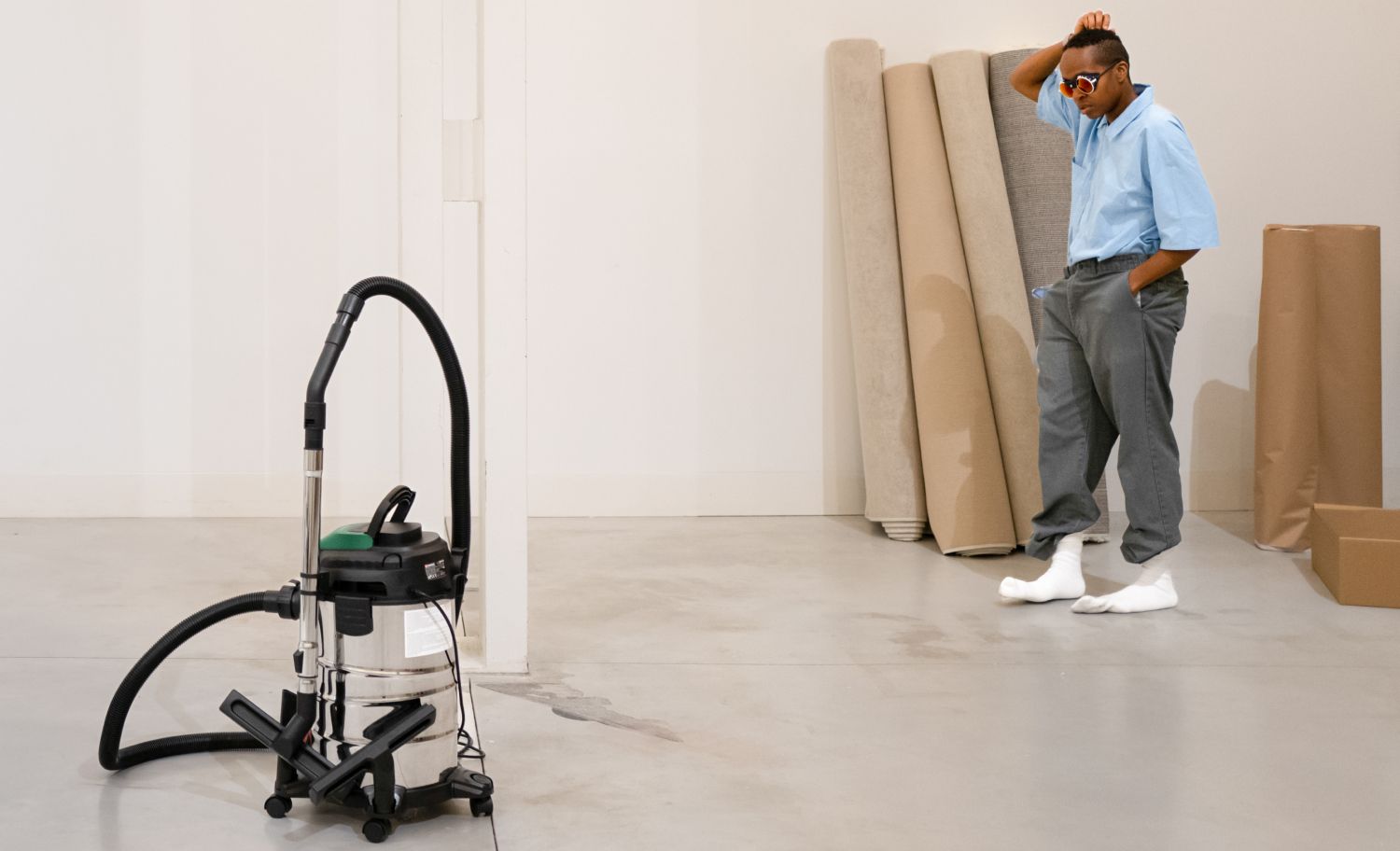 Uma pessoa está em frente a um aspirador, com uma pose confiante, ao lado de tapetes enrolados numa sala vazia. A pessoa está sem sapatos e veste uma camisa azul, calças cinzentas e meias brancas. - Françoise Robert