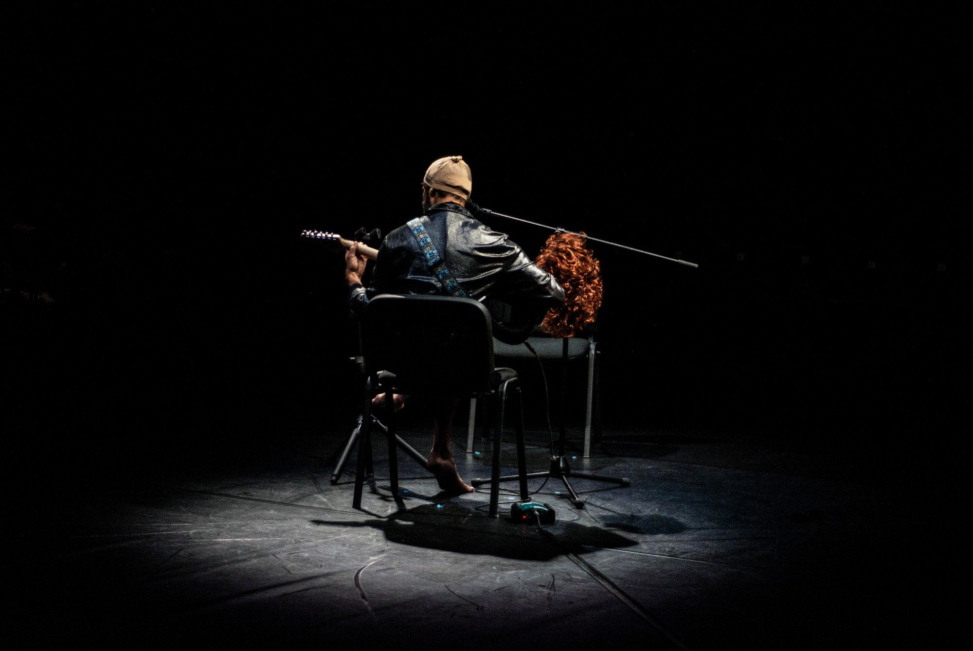 A person on stage playing guitar, viewed from behind, under soft lighting. - Rui Palma