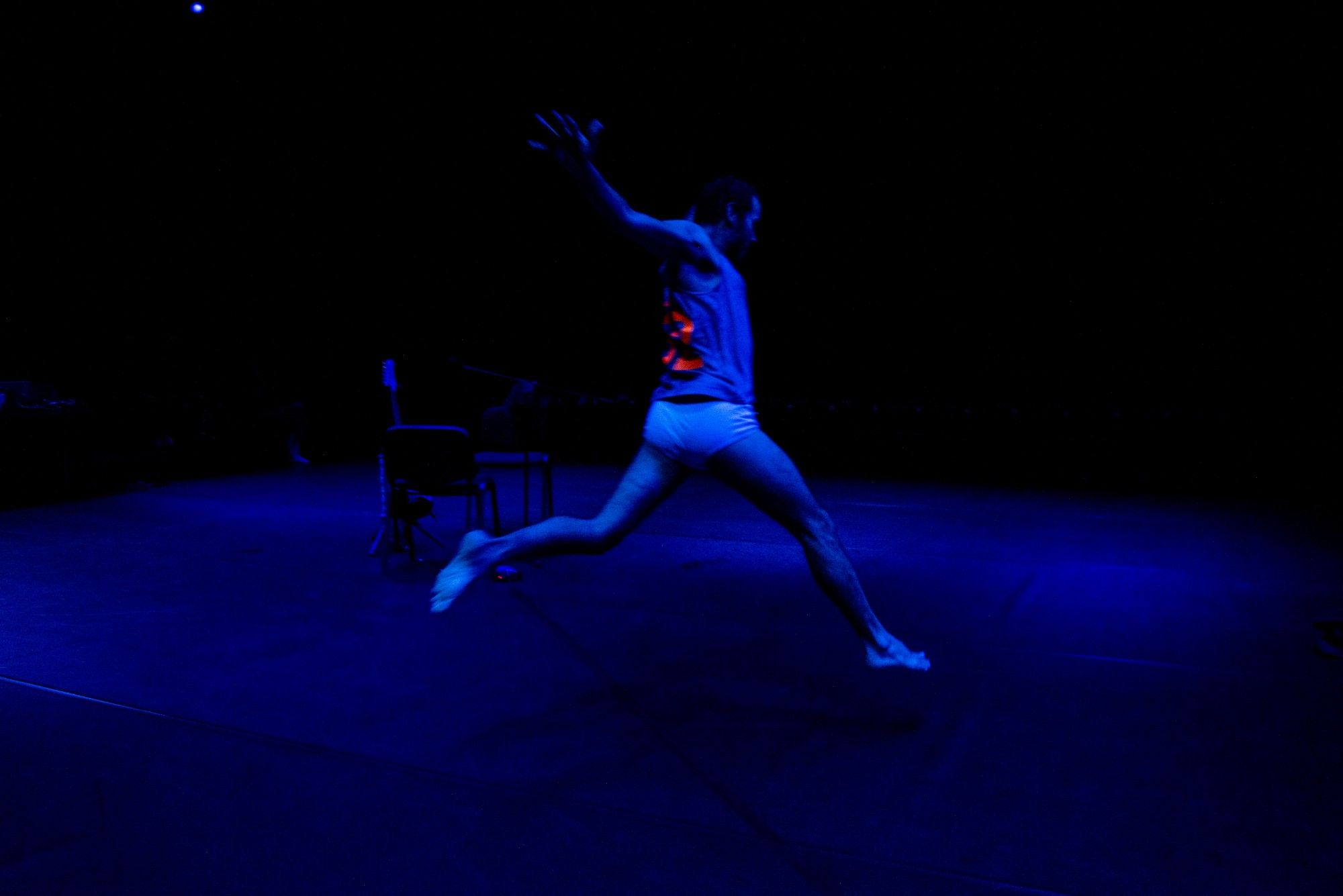 A person in a tank top performing a dance leap on a dark stage, under diffuse blue lighting. - Rui Palma