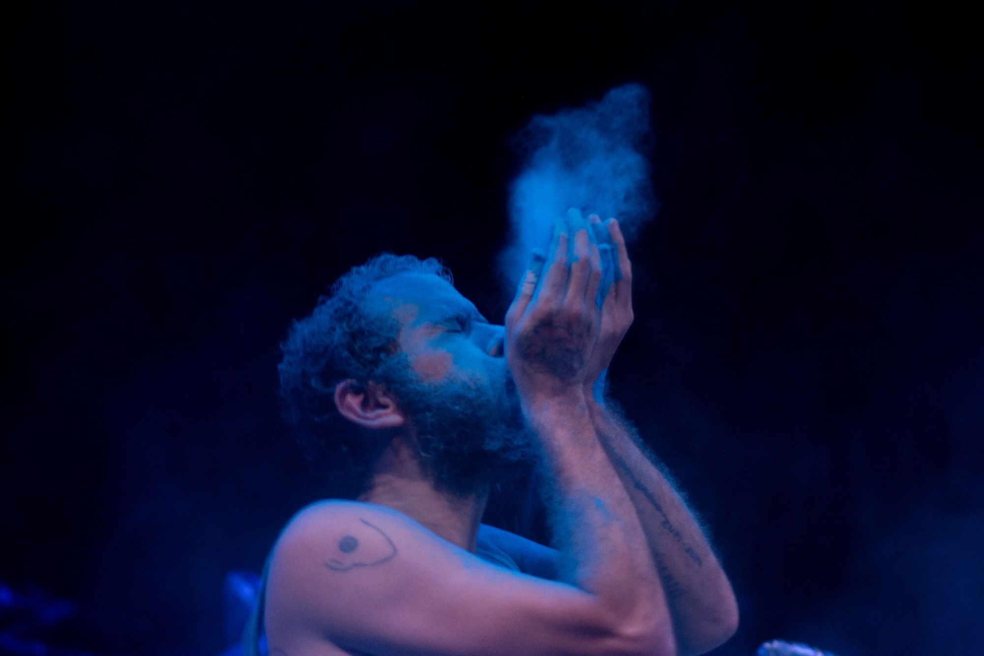 Person with closed eyes and a face painted blue blows powder of the same shade into the air against a dark background. - Rui Palma