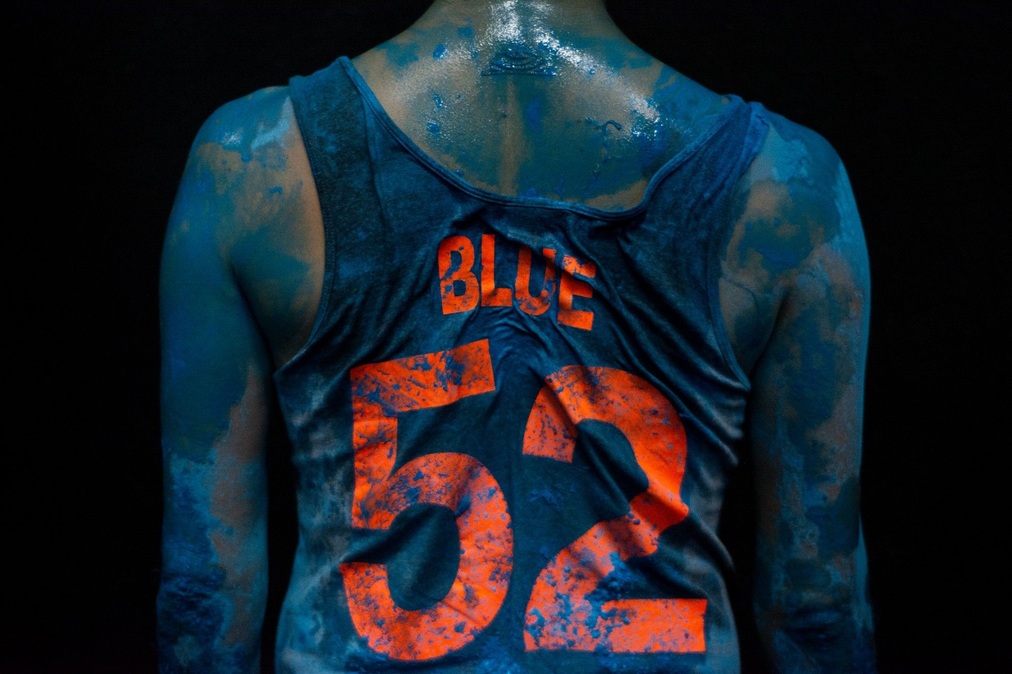 Person with their back turned, blue paint on their skin, and a tank top labeled BLUE 52, against a dark background. - Rui Palma