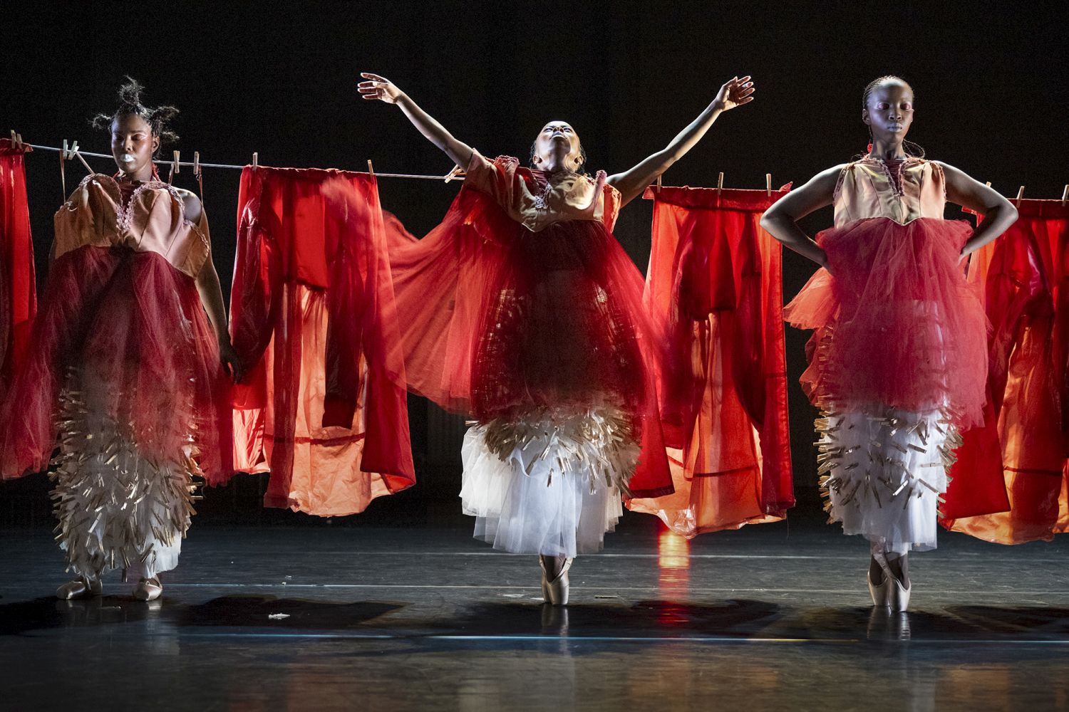 À frente de um estendal com peças de roupa vermelhas estendidas, vemos três corpos que vestem trajes vermelhos e brancos. Todas calçam sapatilhas de ponta de ballet. A pessoa do meio e a pessoa da direita estão em posição de ponta. - Patrick van Vlerken