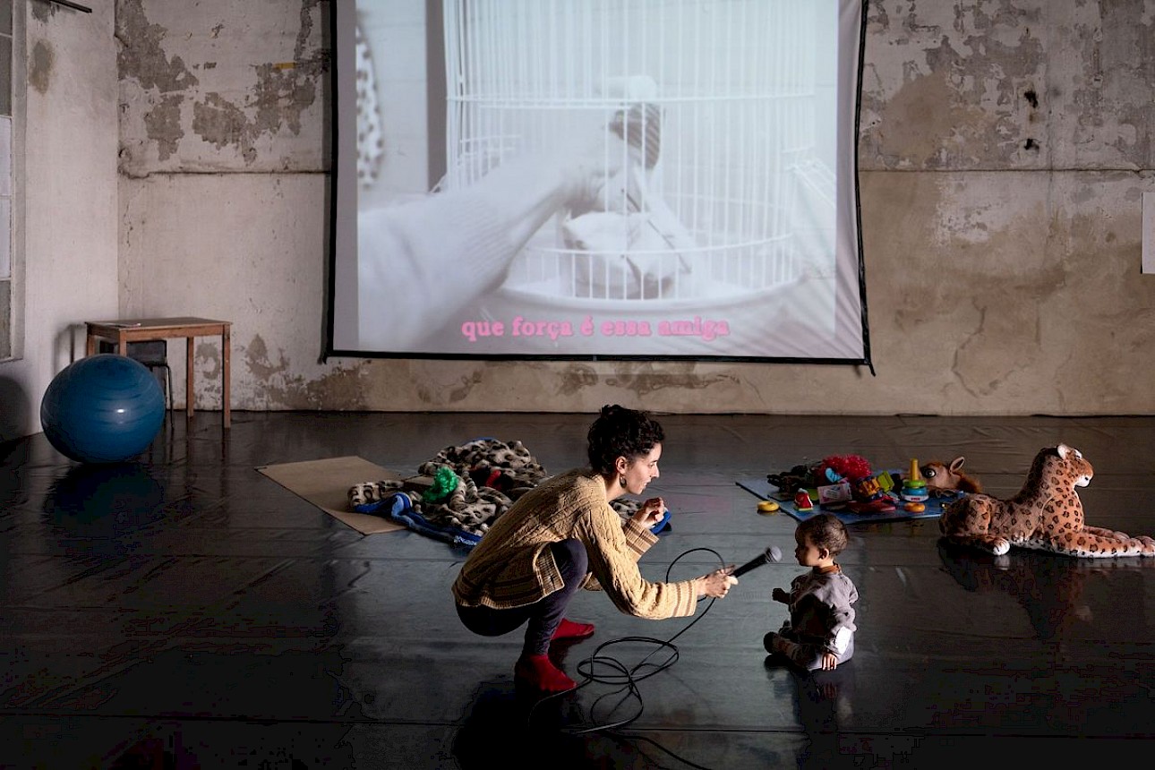 ALKANARA - A person with curly hair kneels with a microphone in front of a child, while a video of a hand and a birdcage plays behind them. - ©Susana Pereira