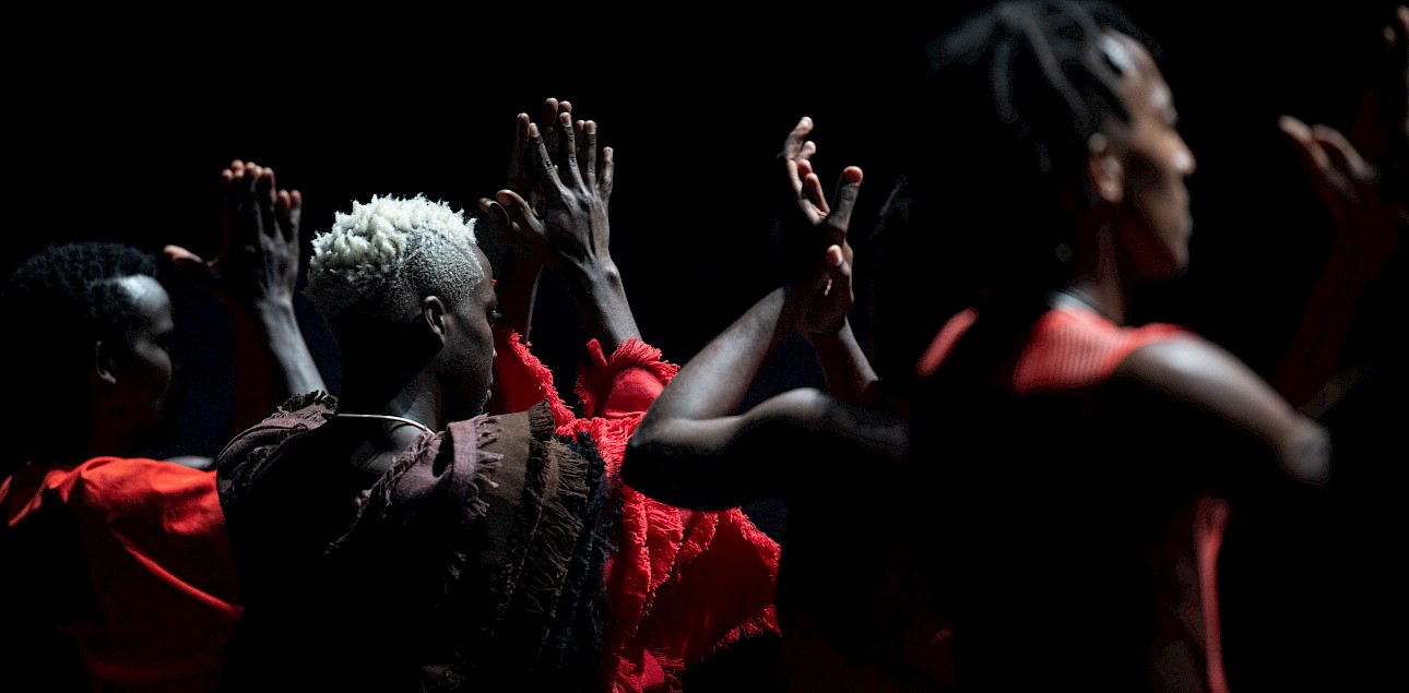 ALKANARA - Several individuals dressed in red clothing raise their hands together. The background is black. - ©Pierre Gondard