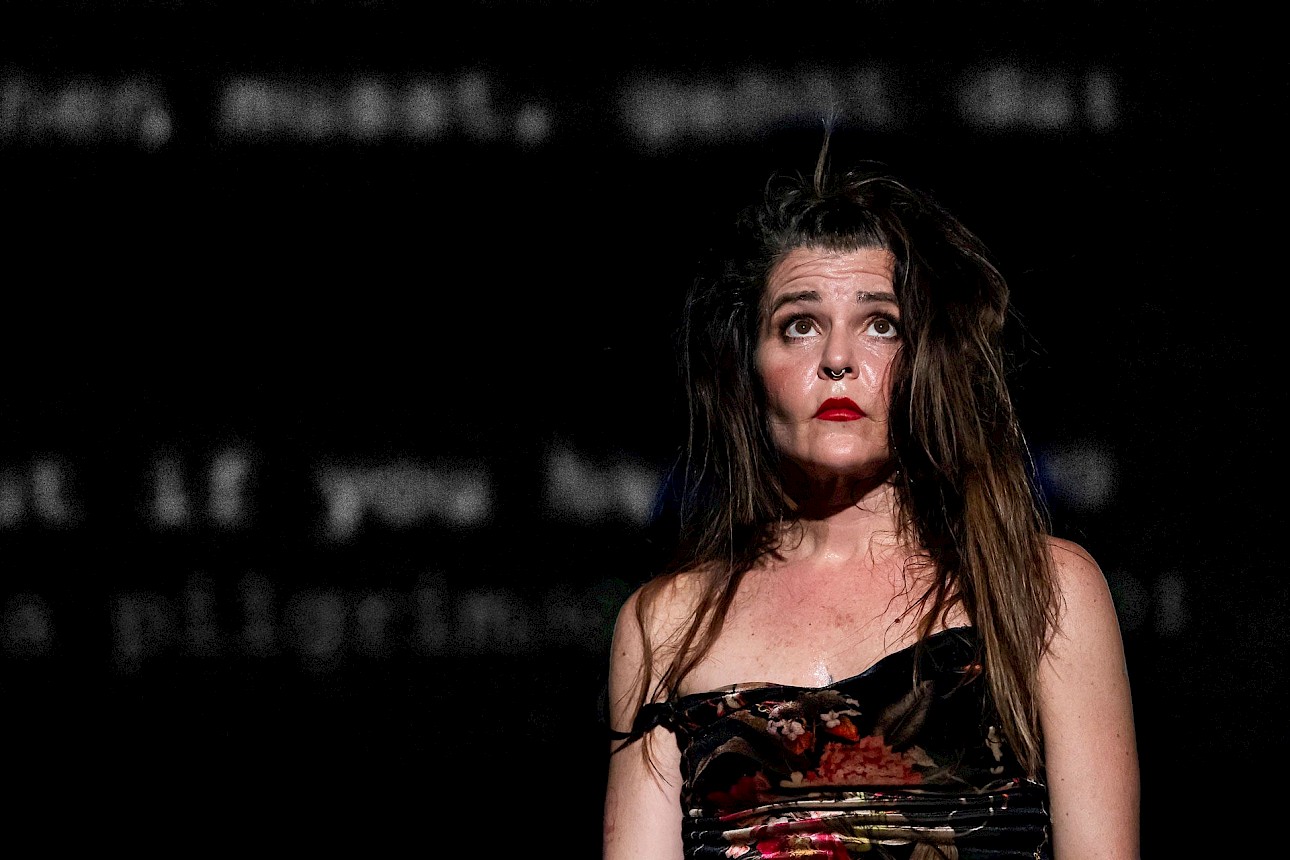 ALKANARA - A caucasian woman with long hair stands in front of a black screen with some unfocused letters. - ©Dajana Lothert