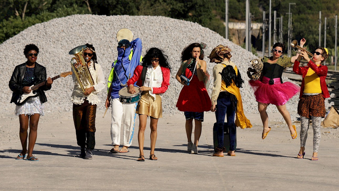 ALKANARA - Um grupo diverso de pessoas com roupas coloridas, alegremente segurando juntos vários instrumentos musicais. - ©Walesca Timmen