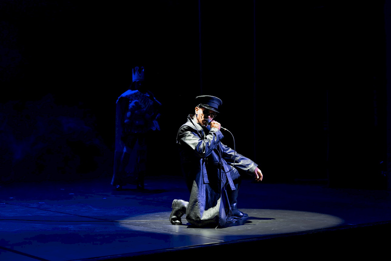 ALKANARA - A performer in a dramatic pose kneels on stage, wearing a leather coat and hat, while holding a microphone against a dark backdrop. - ©Bohumil Kostohryz (TALENTLAB 2025)