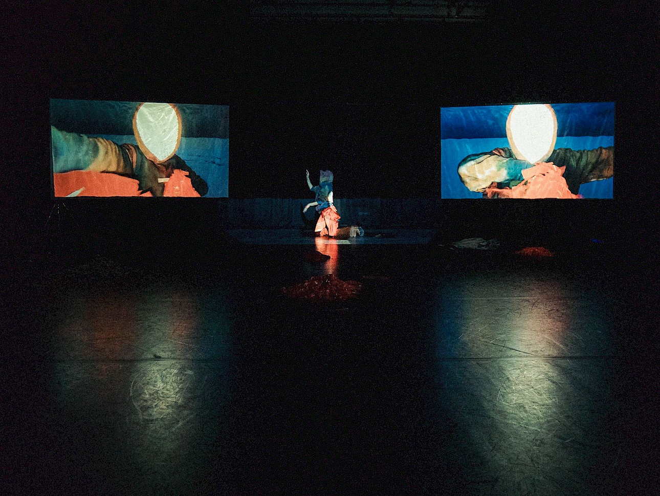 ALKANARA - Uma pessoa vestida com uma roupa colorida com franjas ajoelha-se no palco, rodeada por tecidos esvoaçantes suavemente iluminados num cenário escuro. - ©Gergely Ofner