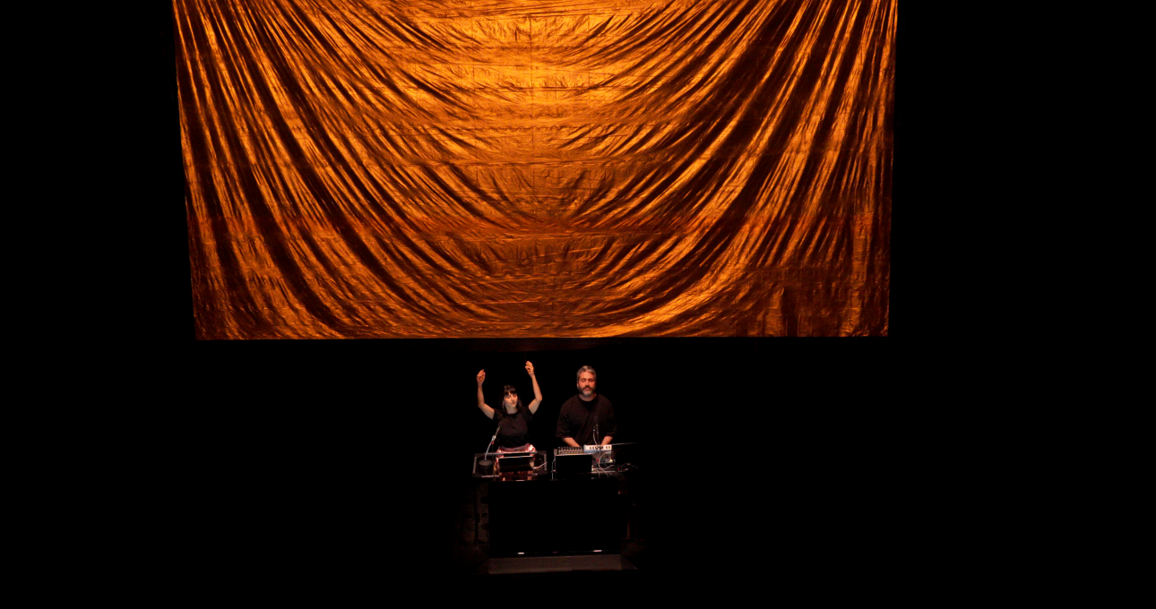 ALKANARA - Duas pessoas atuam no palco, levantando as mãos juntas, num cenário de pouca luz com equipamentos eletrónicos. - ©Firas El Hallak