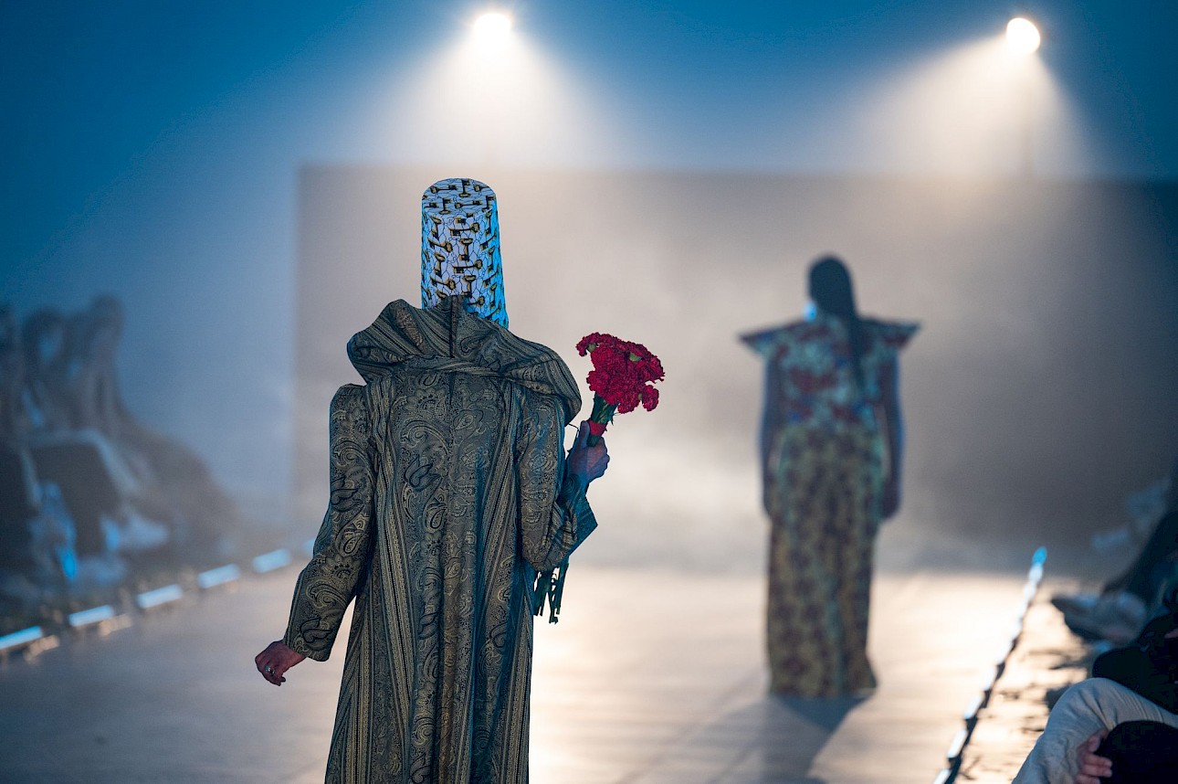 ALKANARA - A person wearing ornate clothing and a patterned hat parades down the catwalk, holding red flowers, with another person in vibrant attire in front of them. - ©José Caldeira