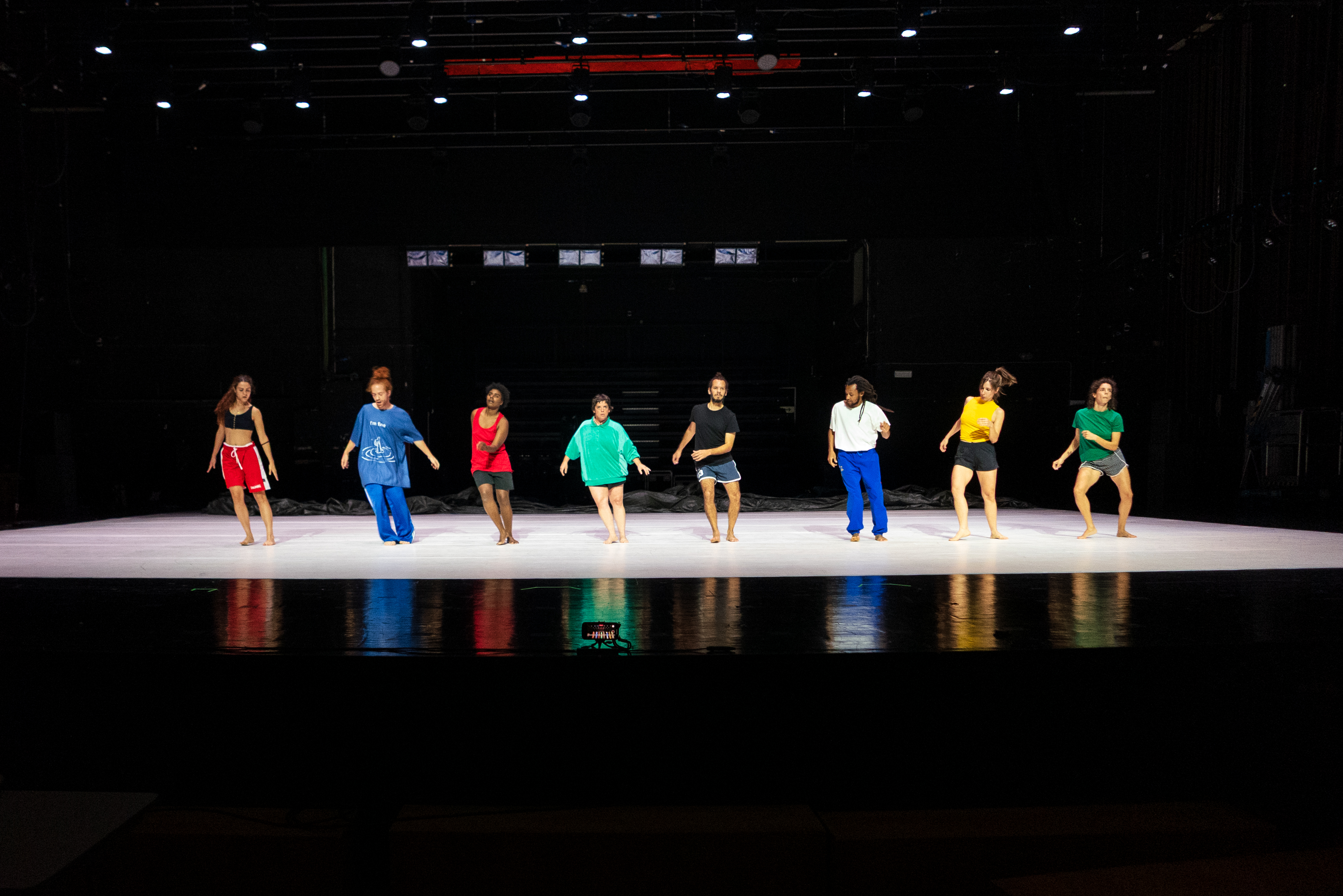 © Walesca Timmen A group of eight dancers performs on stage, wearing colorful outfits. - Walesca Timmen