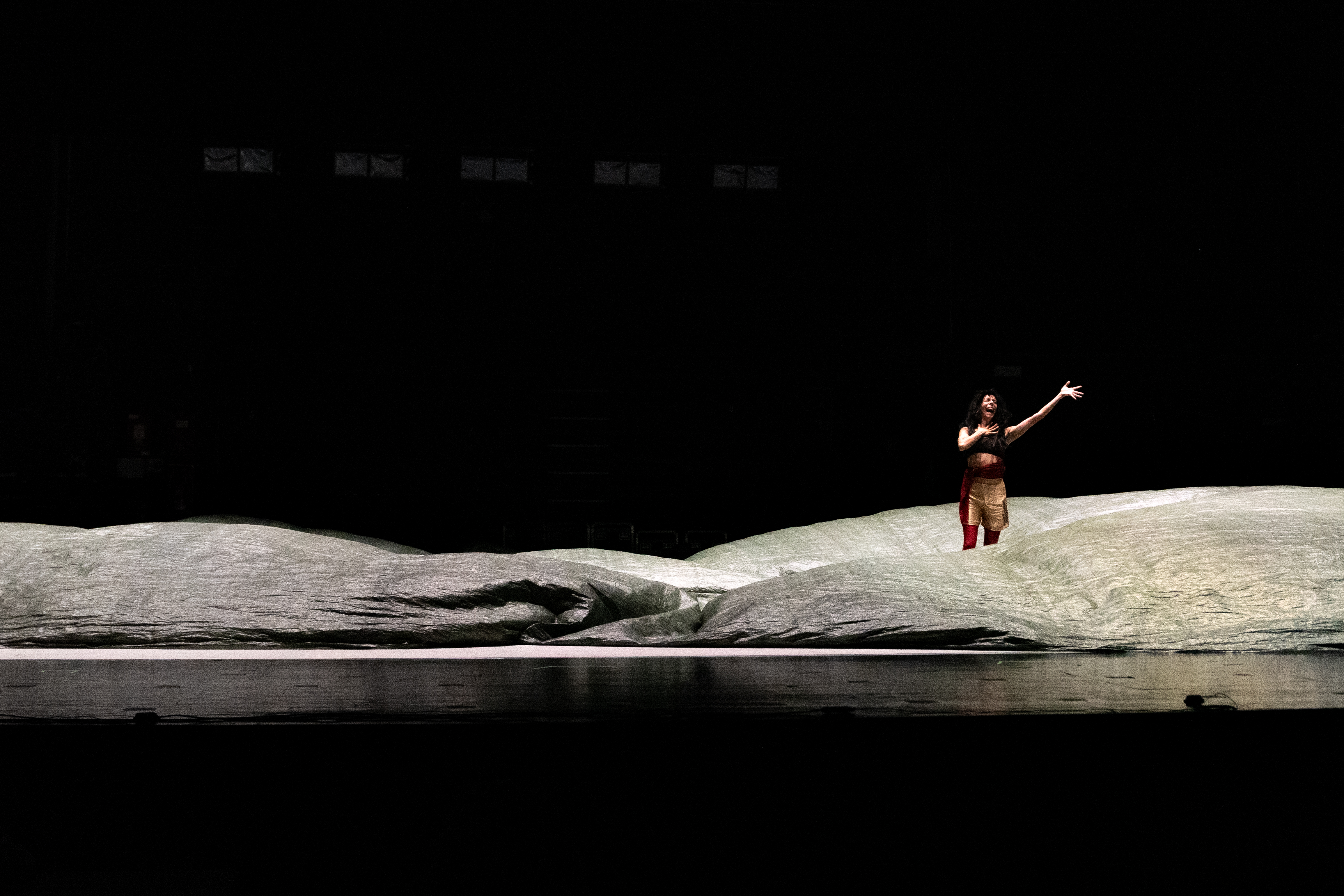 © Walesca Timmen A lone performer stands on a dimly lit stage, surrounded by large, undulating fabric resembling waves. The scene conveys a sense of drama and solitude. - Walesca Timmen