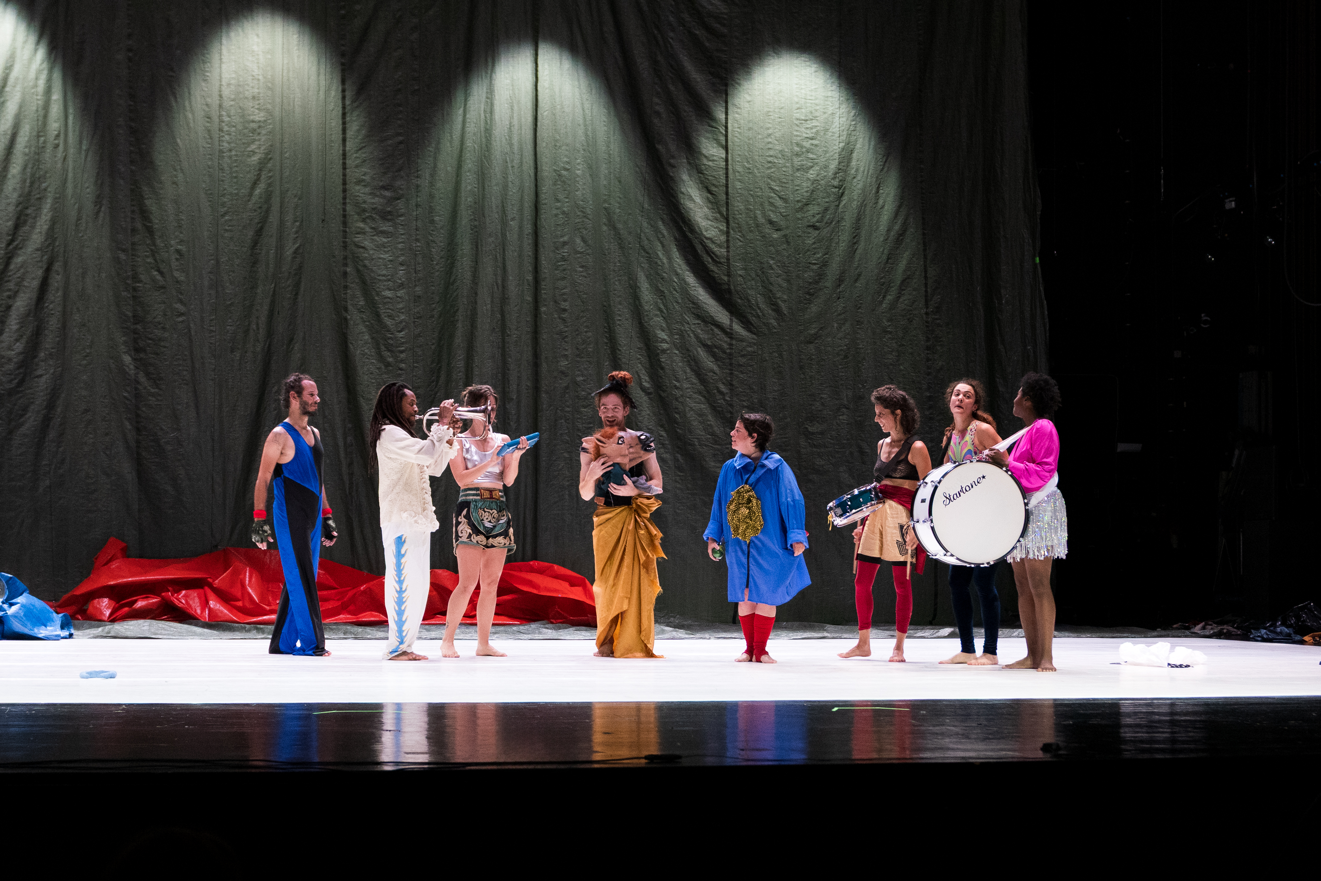 © Walesca Timmen A group of eight people in colorful, eclectic costumes stand on a stage, some holding musical instruments, with a draped backdrop behind them. - Walesca Timmen