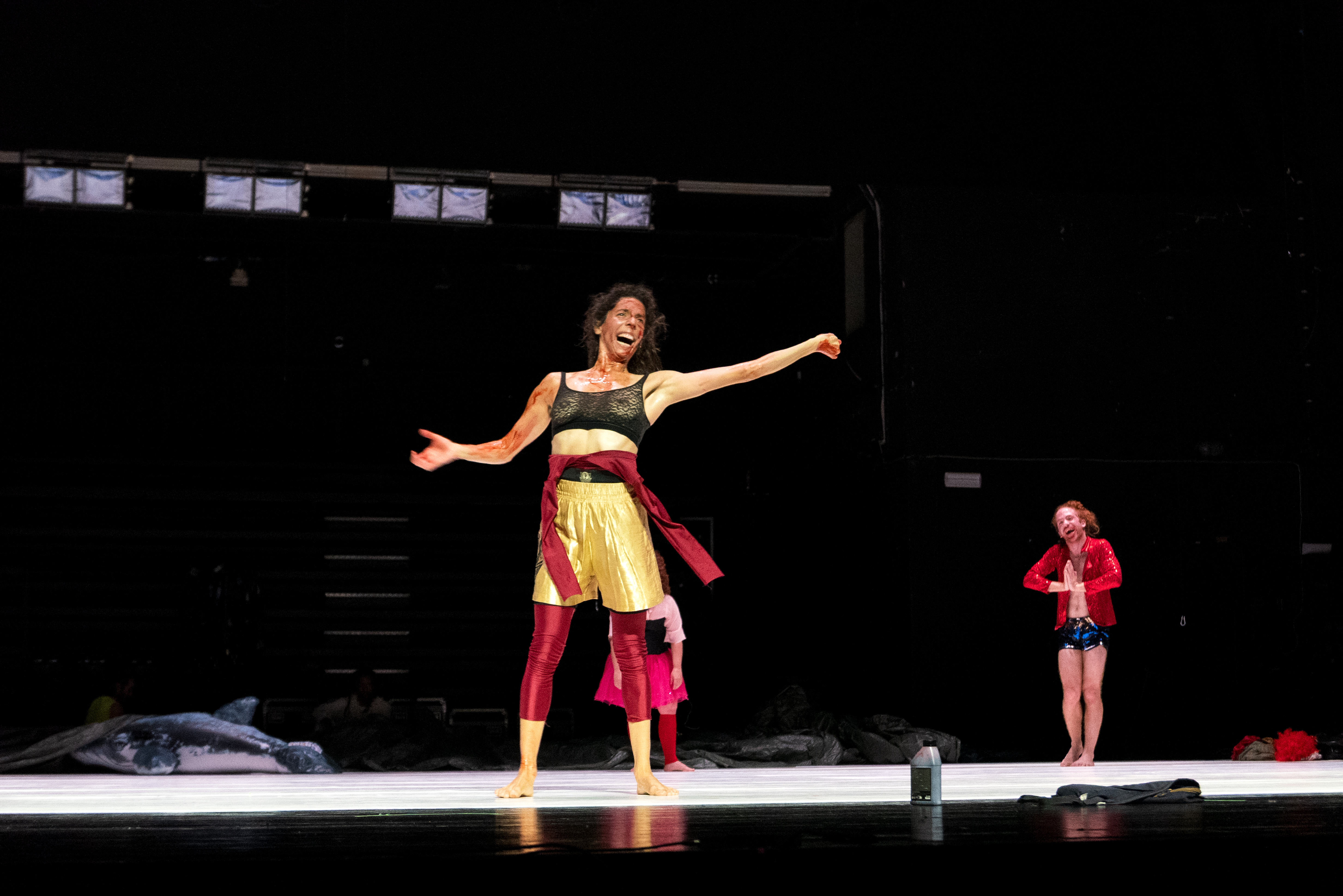 © Walesca Timmen A performer on stage, center, wearing a glittery black top, gold skirt, and red sash, exudes energy and joy. Another dancer in a red jacket smiles in the background. - Walesca Timmen