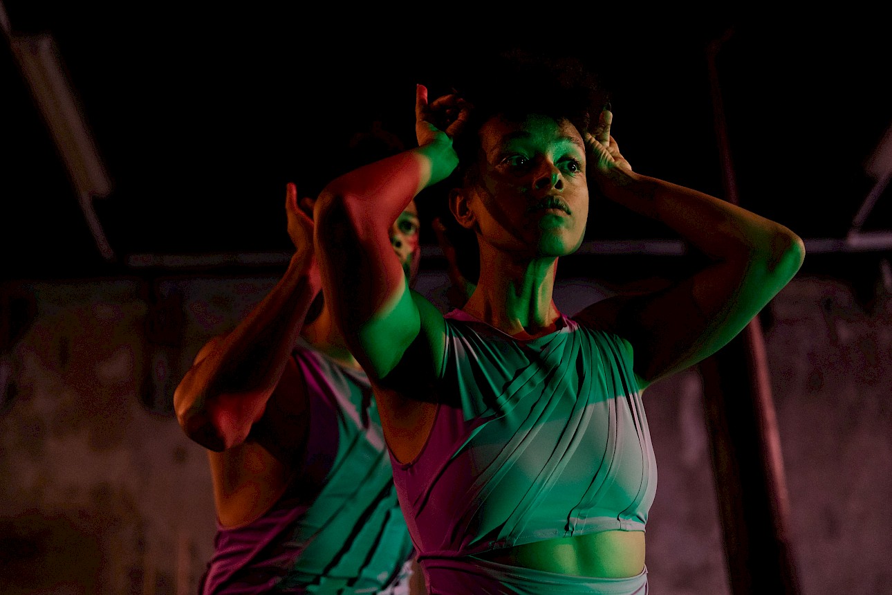 ALKANARA - Two people stretching on a shiny floor, one in a jersey and the other in patterned clothing, with heads down and arms extended forward. - ©Diana Tinoco / Alkantara