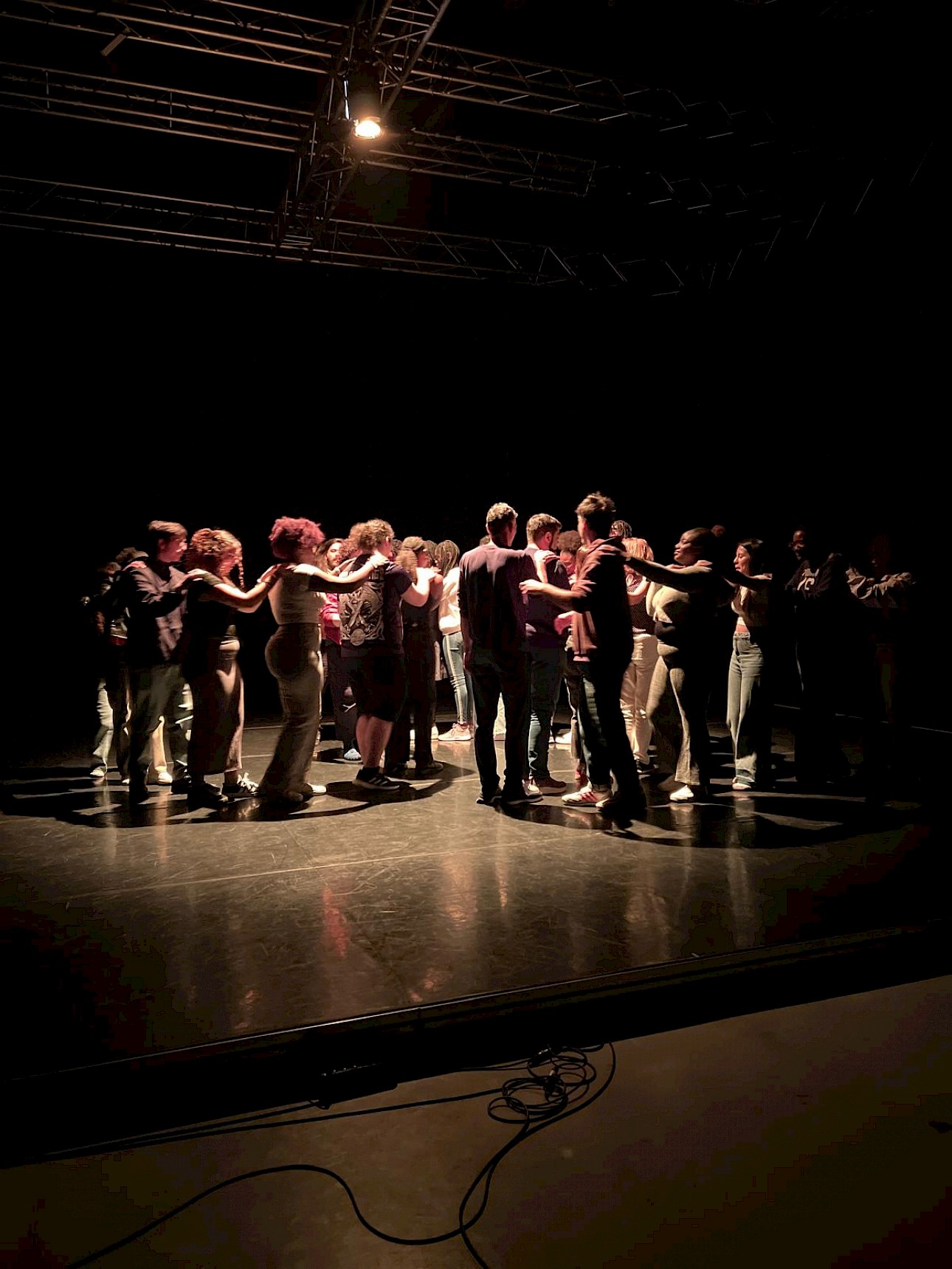ALKANARA - A group of people stands in a cozy circle on a dimly lit stage, arms interlinked, creating a warm, communal atmosphere under soft overhead lighting. - ©Aurora