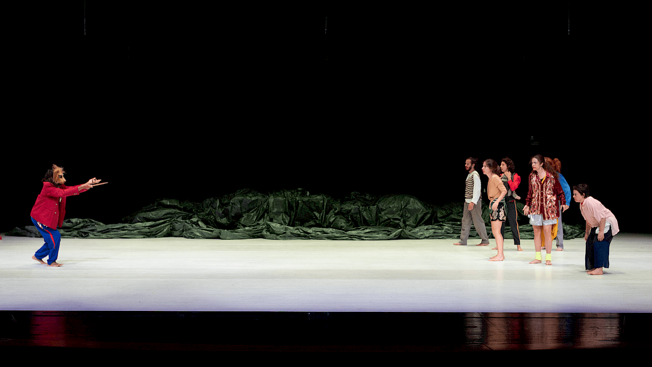 ALKANARA - A performer in a red jacket gestures toward a group of seven people on stage. They're barefoot, standing on a bright surface with a dark backdrop. - ©Walesca Timmen