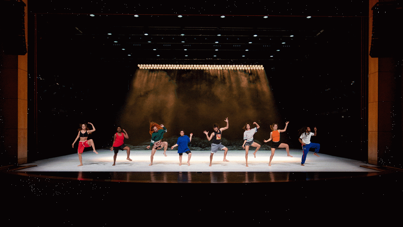 ALKANARA - Uma pessoa intérprete, de casaco vermelho, faz um gesto em direção a um grupo de sete pessoas em palco. Estão descalças, sobre uma superfície clara, com um fundo escuro. - ©Walesca Timmen
