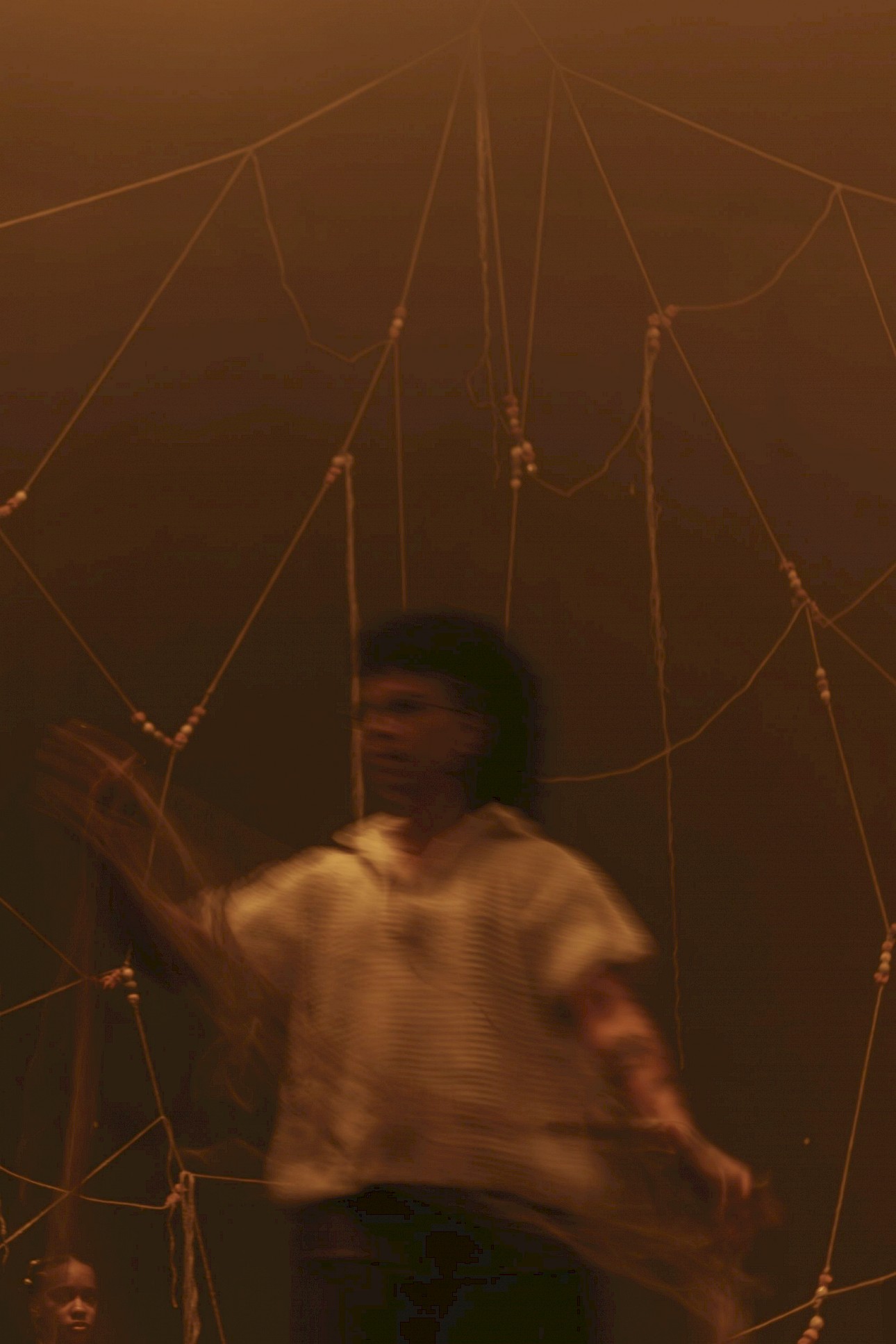 ALKANARA - Uma pessoa em movimento, desfocada, destaca-se contra um fundo de cordas entrecruzadas que criam padrões geométricos. - ©Letícia Diniz