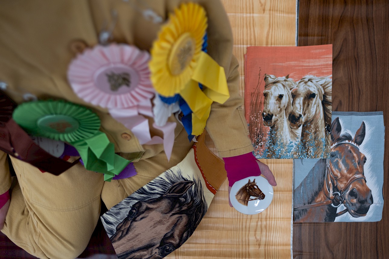 ALKANTARA - Person adorned with colorful horse show ribbons holds a horse-themed plate. Nearby are equestrian paintings and a horse-patterned pillow. - ©Raquel Melgue