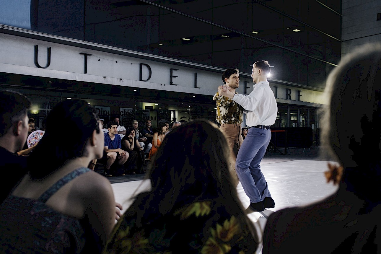 ALKANTARA - Dois homens a dançar a par num palco ao ar livre, à noite, vistos por trás de um público desfocado. Ao fundo, destaca-se um edifício moderno de vidro. - ©Alice Brazzit