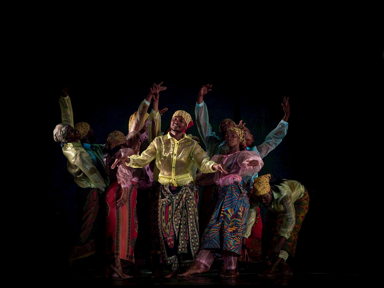 ALKANTARA - Um grupo dança com roupas coloridas, de padrões e lenços na cabeça, executando uma coreografia contra um fundo escuro. - ©Andreas Hagenbach