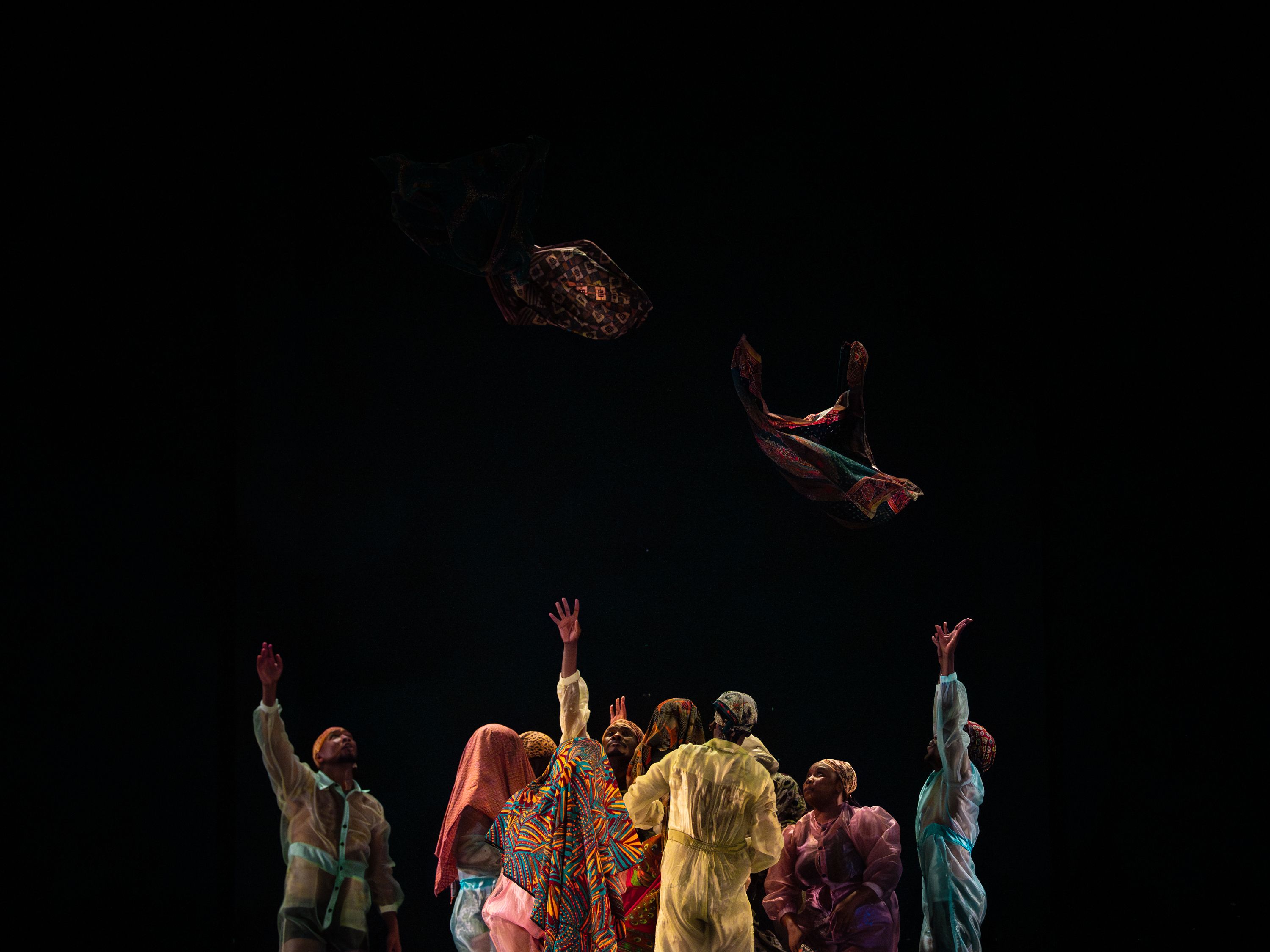 © Andreas Hagenbach Um grupo de pessoas com roupas coloridas está em círculo, estendendo os braços para cima, enquanto pedaços de tecido com padrões flutuam acima de si contra um fundo escuro. - Andreas Hagenbach