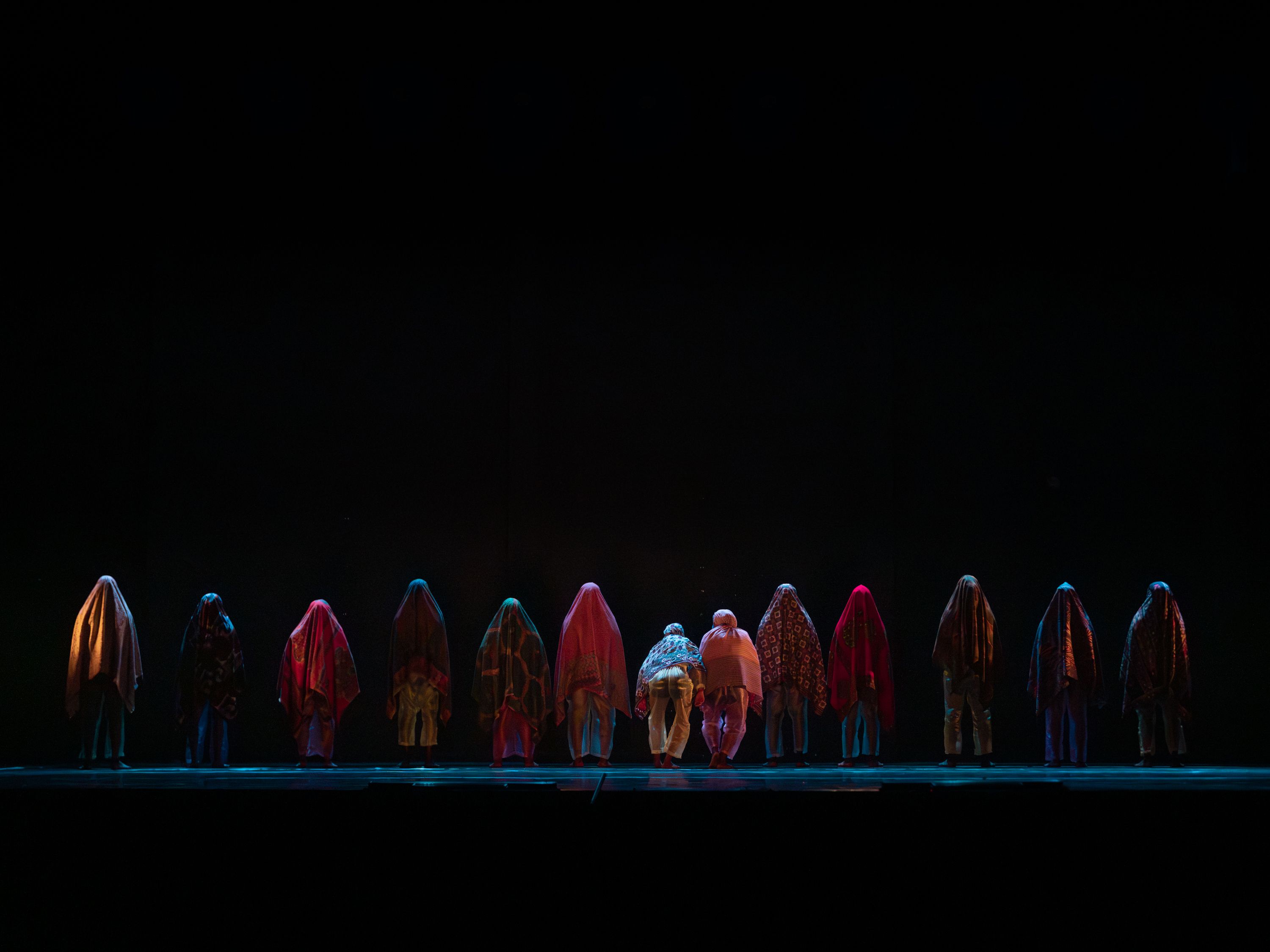 © Andreas Hagenbach Uma linha de pessoas cobertas por tecidos coloridos e de padrões está posicionada num palco contra um vasto fundo escuro. - Andreas Hagenbach