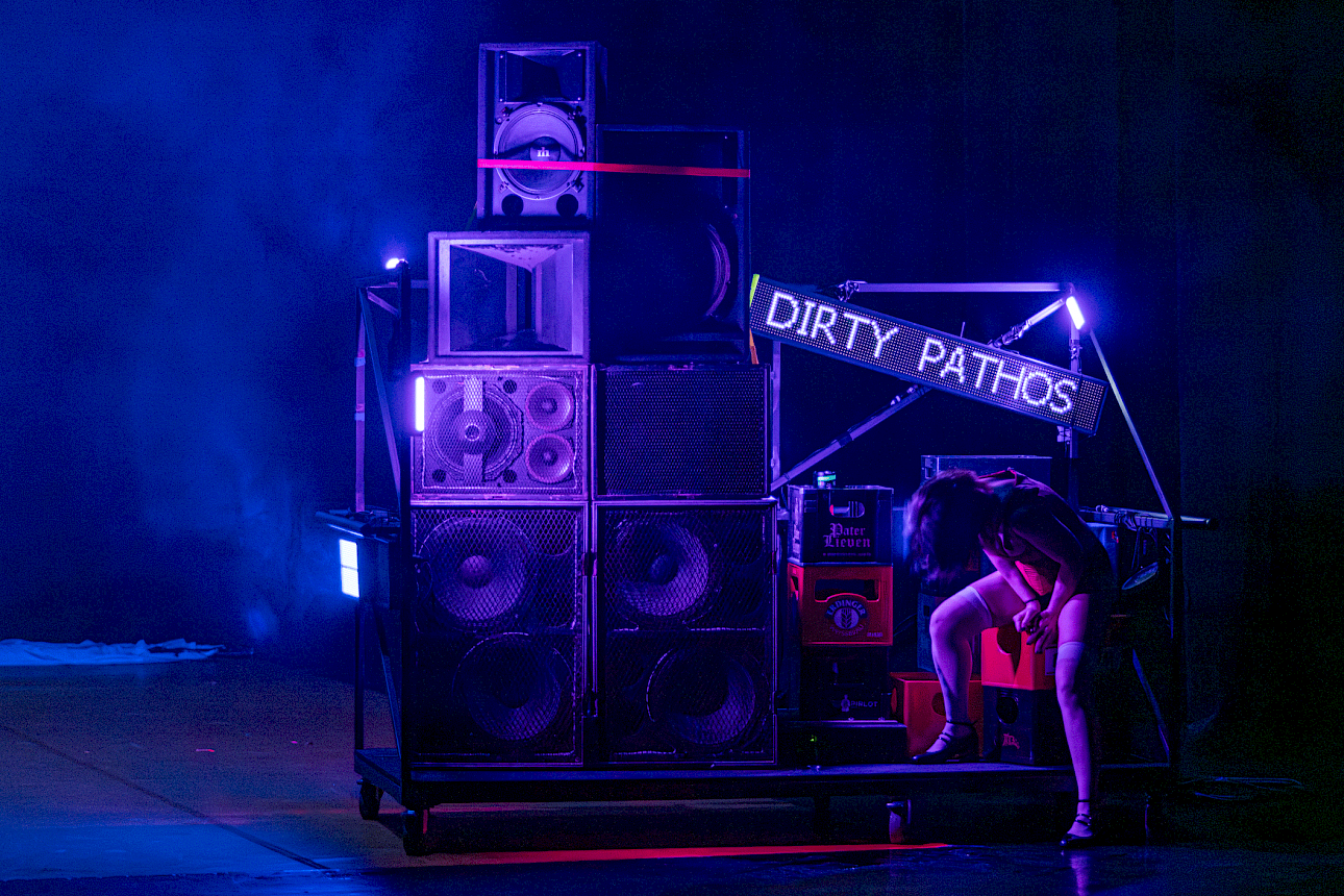 ALKANTARA - Uma performer sentada numa estrutura móvel ao lado de uma grande pilha de colunas de som, sob uma iluminação cénica azul e roxa. Um letreiro eletrónico aceso exibe o texto "DIRTY PATHOS". - ©Mayra Azzi