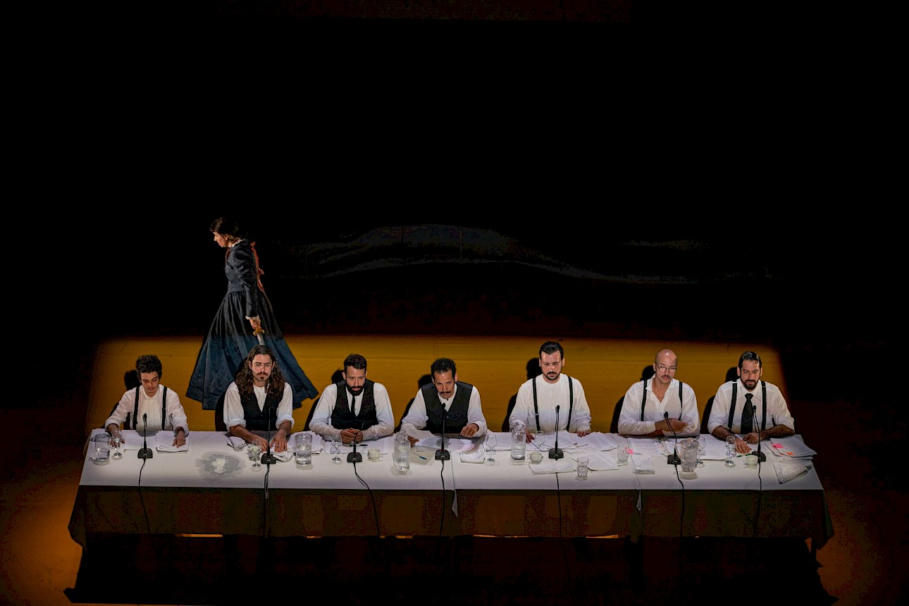 ALKANTARA - Sete homens com camisas brancas e coletes ou suspensórios escuros estão sentados alinhados atrás de uma longa mesa comprida num palco iluminado. À frente de cada um há microfones e papéis espalhados. Ao fundo, na penumbra, uma figura com um vestido longo e escuro caminha de perfil. - ©Mayra Azzi