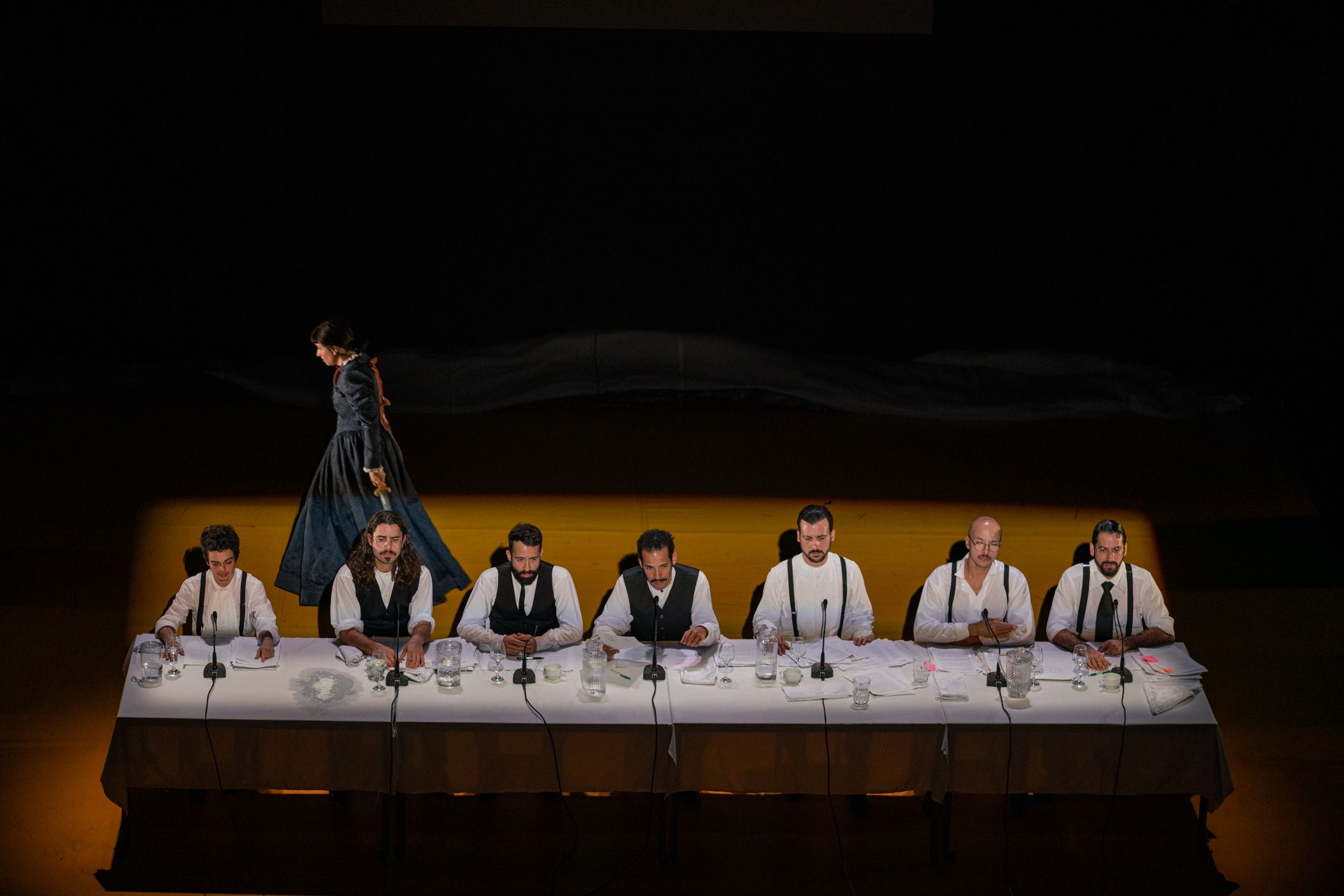 © Mayra Azzi Sete homens com camisas brancas e coletes ou suspensórios escuros estão sentados alinhados atrás de uma longa mesa comprida num palco iluminado. À frente de cada um há microfones e papéis espalhados. Ao fundo, na penumbra, uma figura com um vestido longo e escuro caminha de perfil. - Mayra Azzi