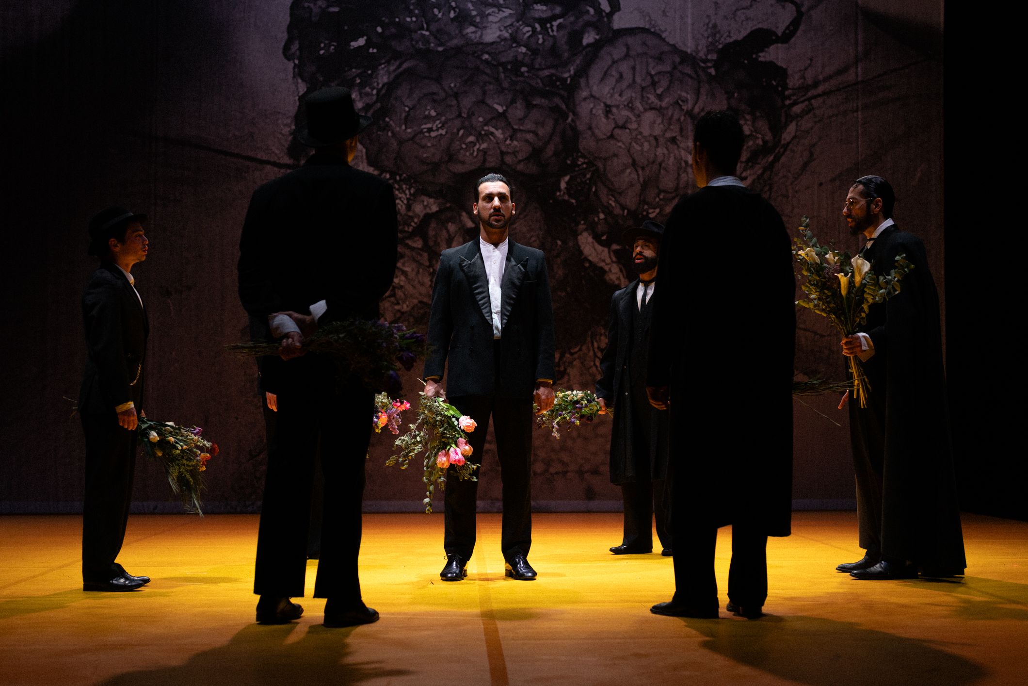 © Mayra Azzi Um grupo de homens vestidos com fatos escuros forma um círculo num palco iluminado a amarelo. O homem ao centro está virado para a frente, enquanto os outros seguram ramos de flores. Ao fundo, destaca-se um grande painel texturado com uma imagem abstrata escura. - Mayra Azzi