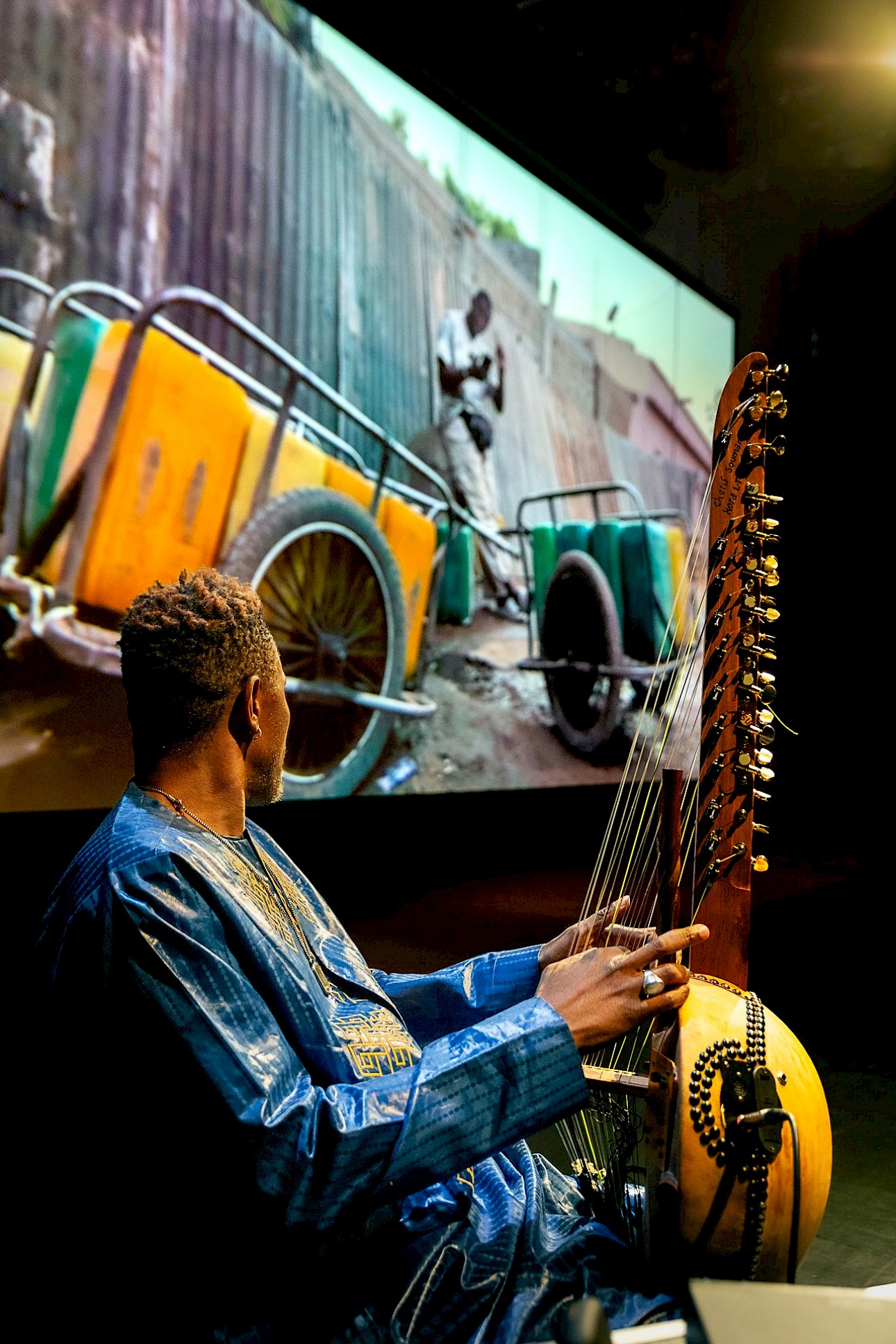 ALKANTARA - A musician seen from behind, wearing a shiny blue robe, playing a kora on stage. In the background, a large screen projects an image of a cart with yellow jerrycans on a street. - ©Claudia Ndebele