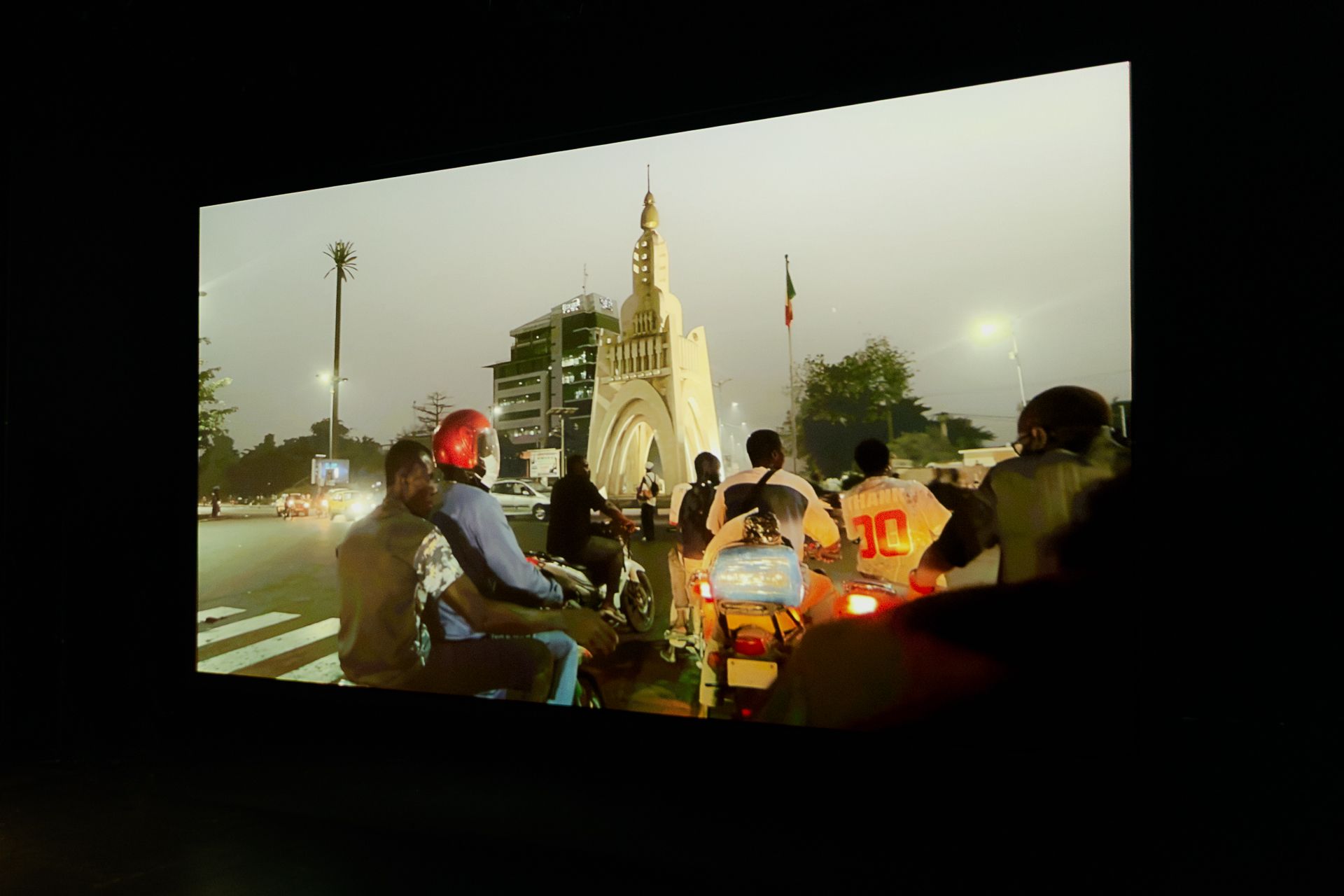 A large screen on a dark stage projects a city scene at dusk, showing several people on motorbikes riding down an avenue towards a large white monument. - Claudia Ndebele