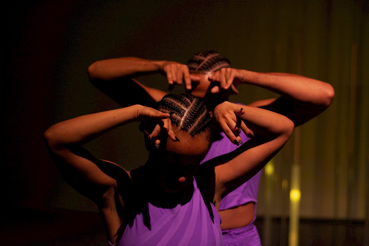ALKANTARA - Close-up of two people with braided hair under dramatic orange lighting. Both have their hands raised near their heads, with fingers positioned expressively, casting shadows over their faces. - ©Rui Palma