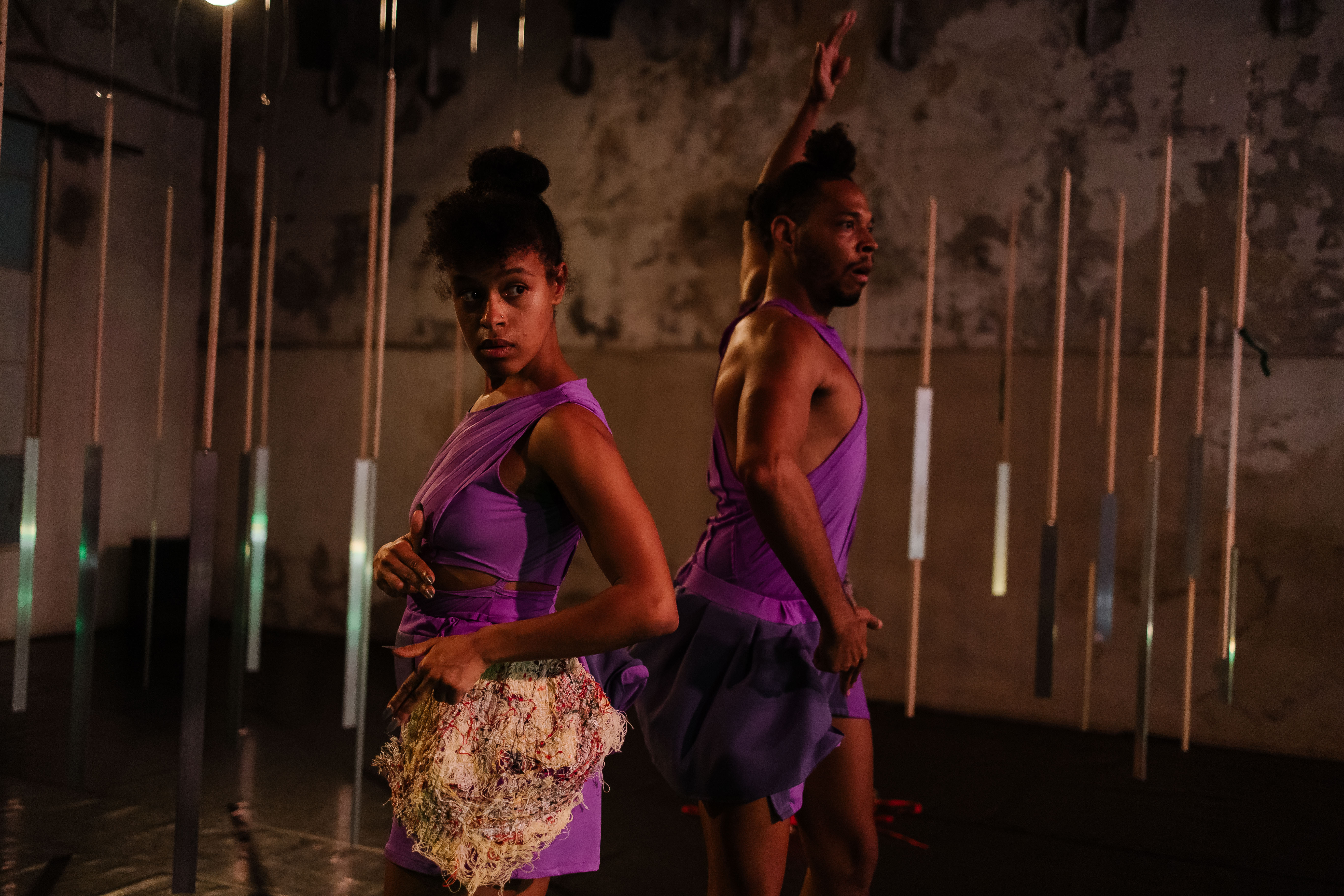 Two performers on a dimly lit stage, wearing coordinating purple outfits. The person in the foreground, with their hair in a high bun, looks intensely to the left, holding a textured, light-coloured piece of fabric at their hip; the person behind has one arm raised, looking to the right. Thin vertical rods hang in the background. - Diana Tinoco