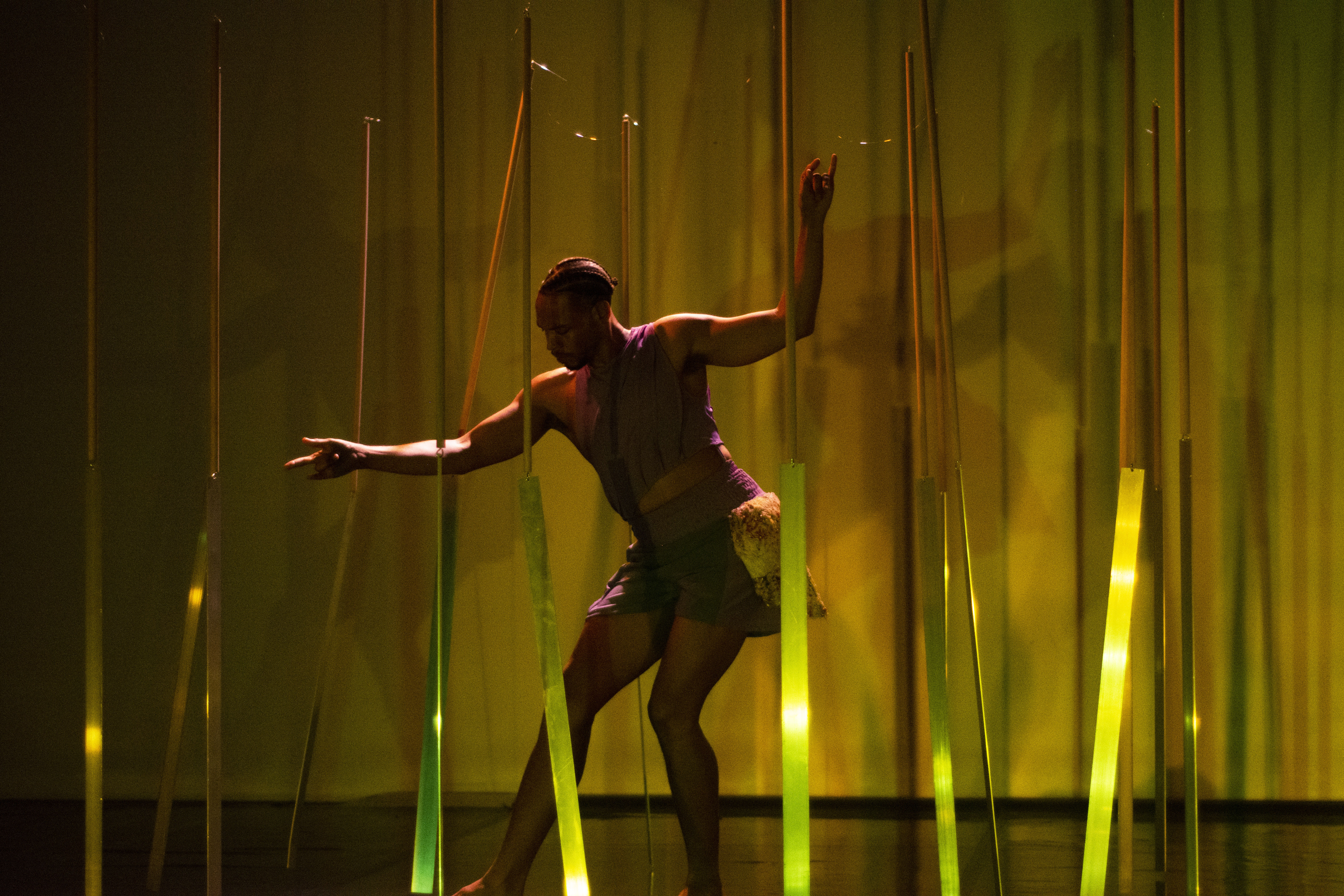 A performer with braided hair dances on a stage, bending their knees and stretching out their arms. They move among several vertical strips that glow with green and yellow light, set against a warm background. - Rui Palma