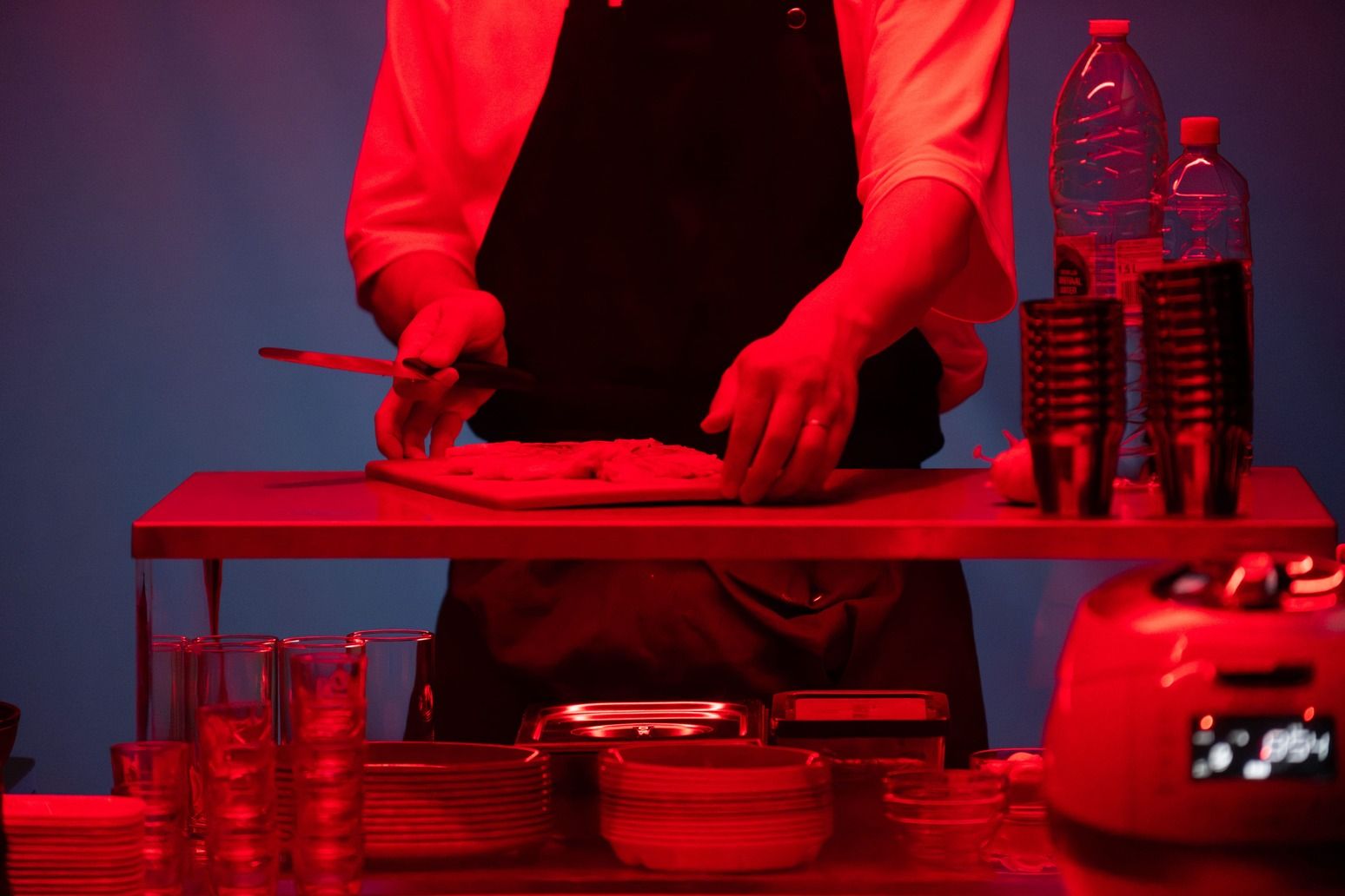 © Bea Borgers A person wearing a white t-shirt and black apron holds a knife and cuts food on a chopping board at a counter. The entire scene is lit by a strong red light against a blue background. - Bea Borgers