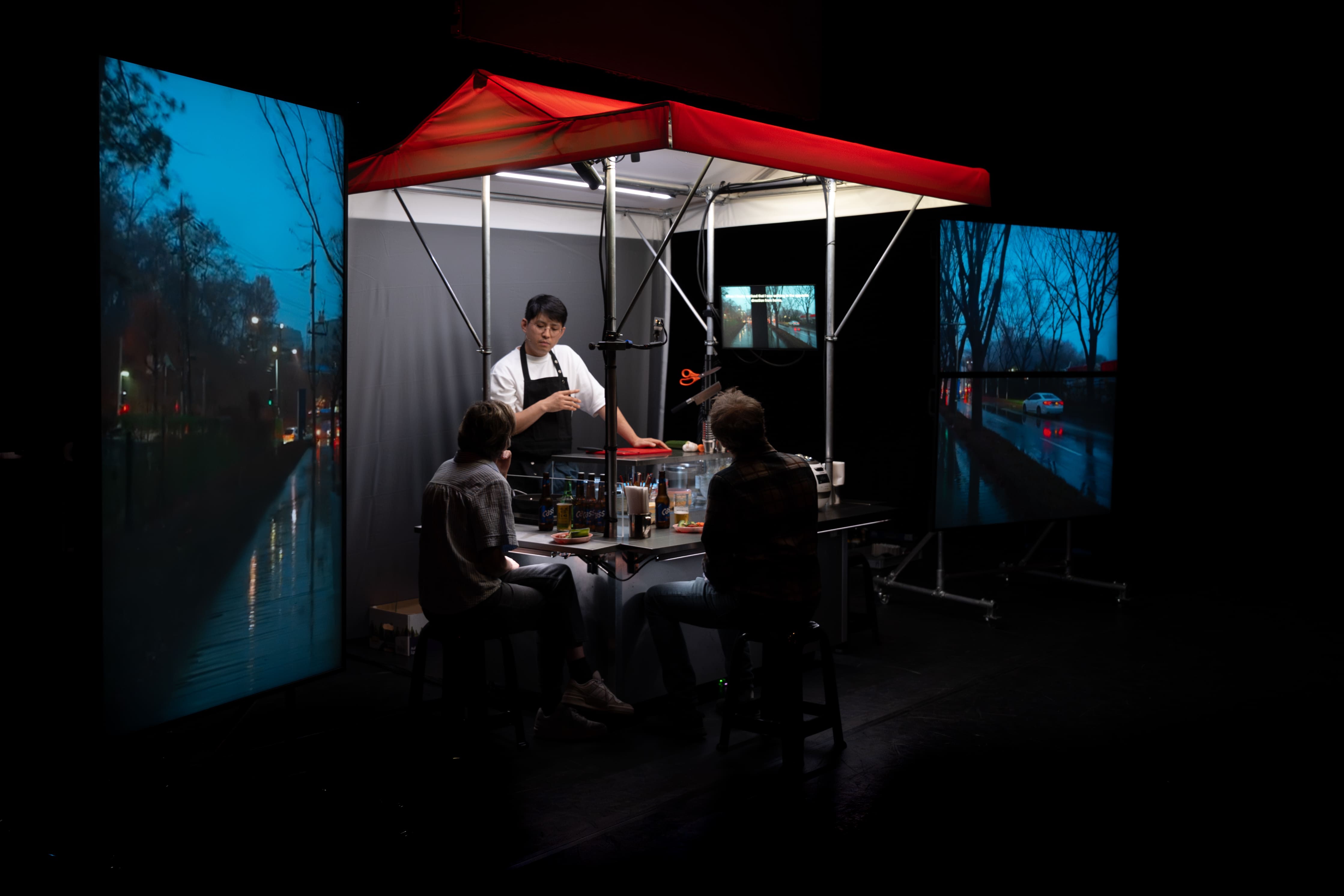 © Bea Borgers A closer view of a stage art installation, focused on a Korean food stall under a red awning. A chef in a black apron talks to two customers seated at the counter. The stall is framed by two large vertical LED screens displaying rainy, night-time cityscapes, reflecting the city lights. - Bea Borgers